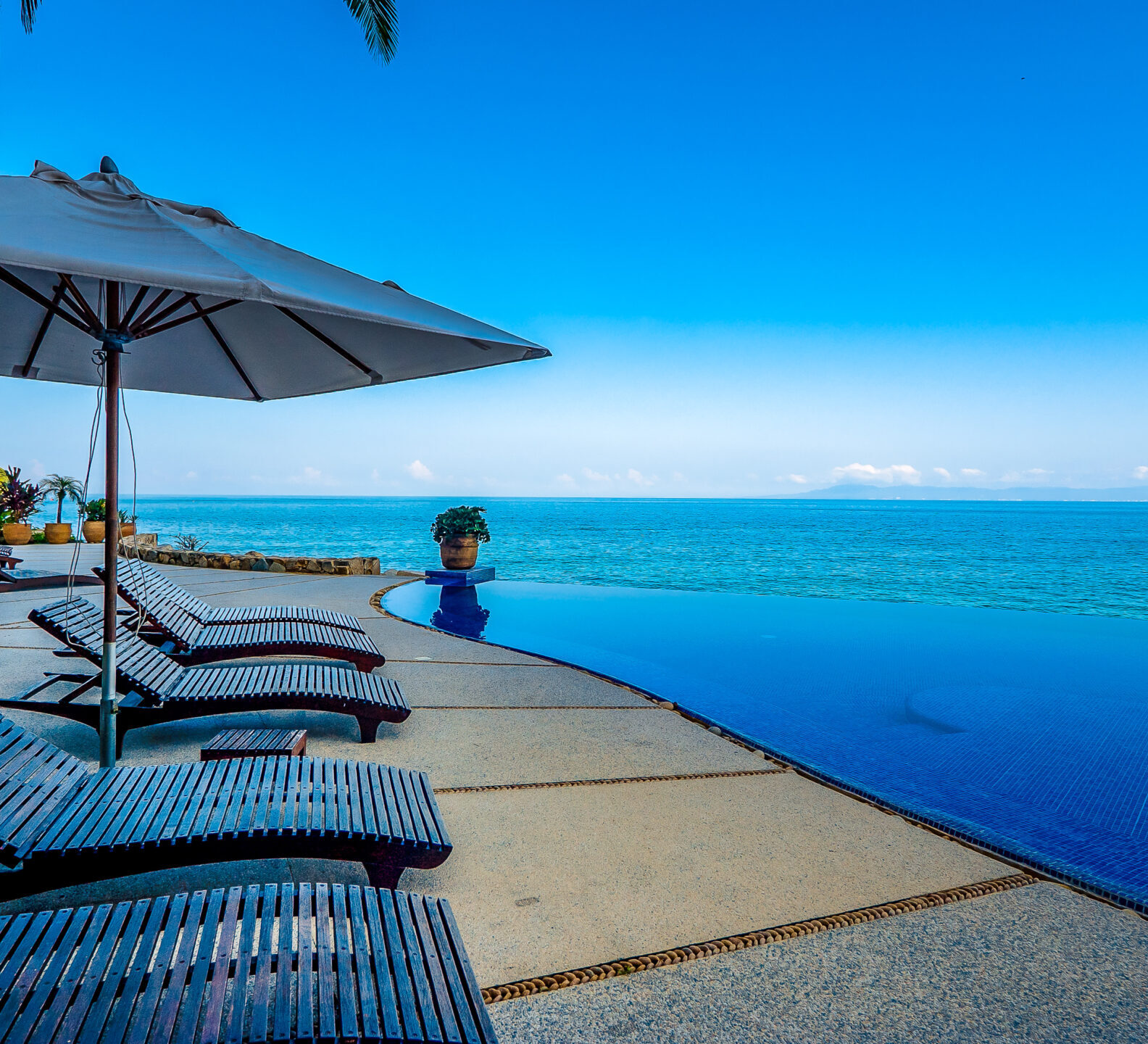 Poolside lounge chairs beneath umbrellas beside an infinity pool overlooking the ocean.