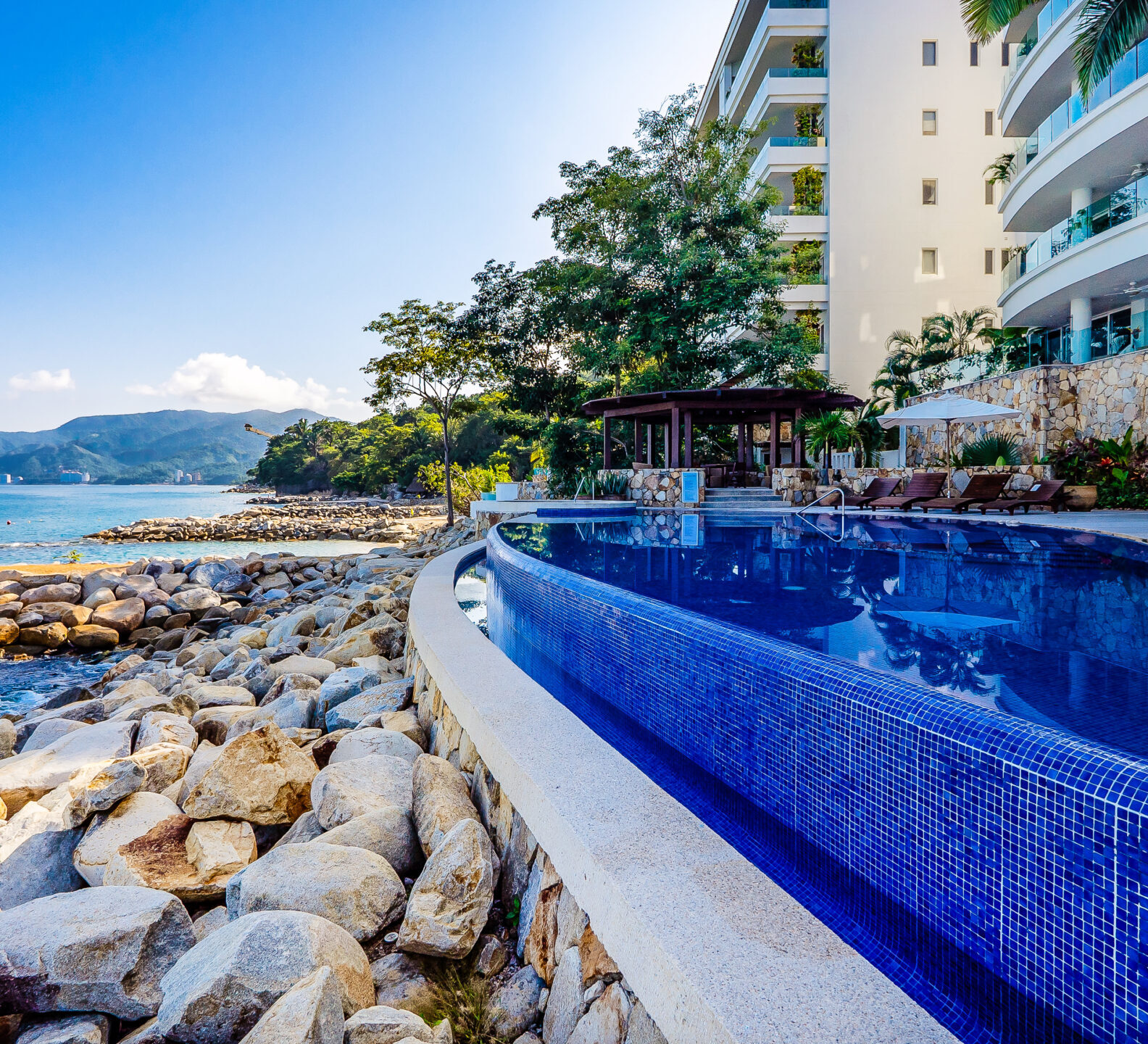Infinity pool beside a rocky shoreline with condominium buildings in the background.