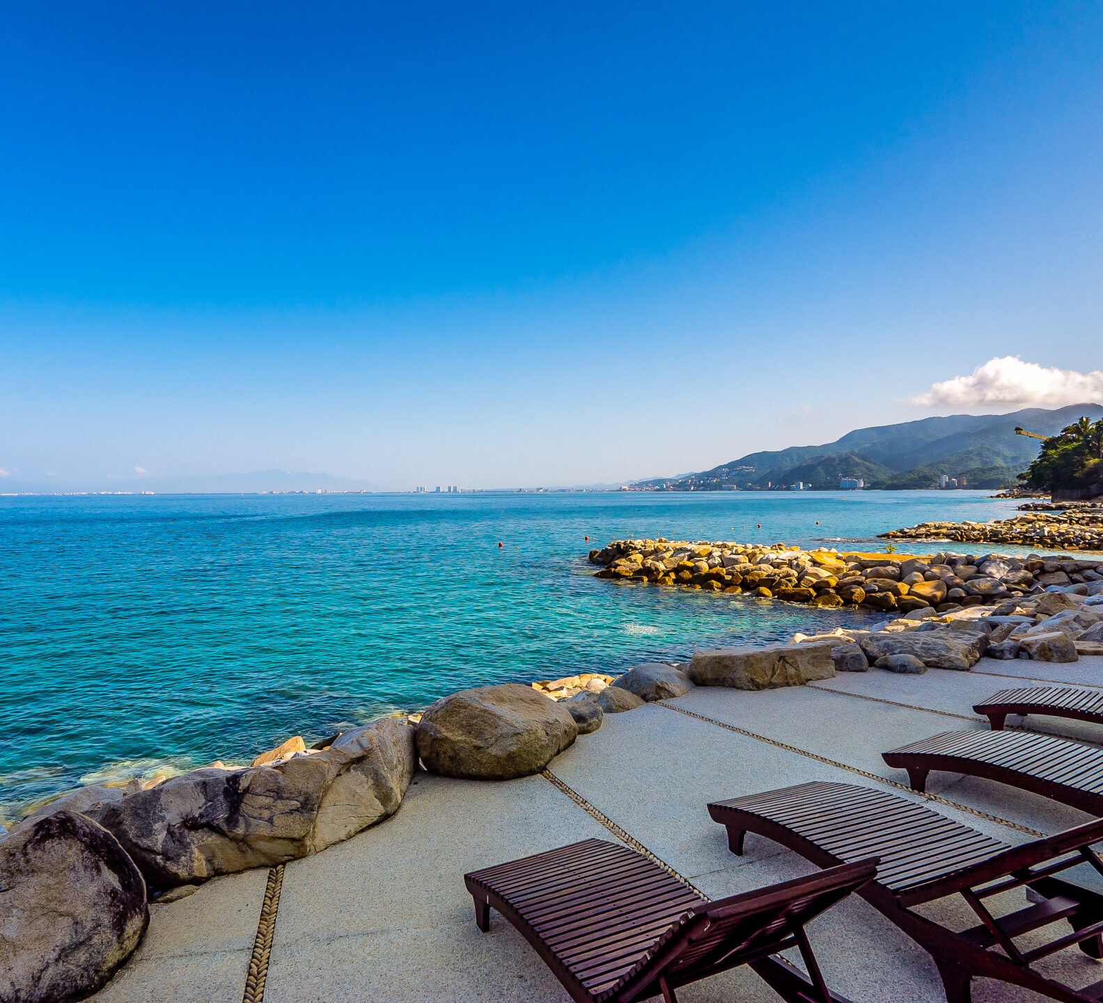 Lounge chairs arranged beside the ocean with rocky barriers and distant mountain views.