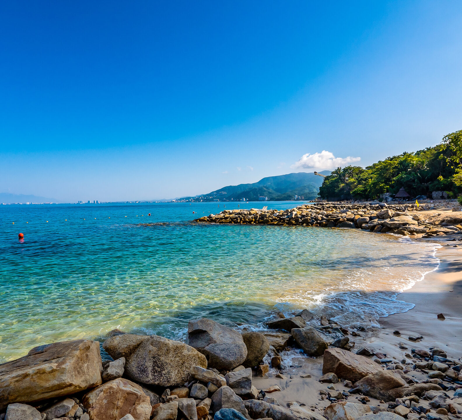 Rocky shoreline with clear blue water and distant mountains beneath a bright sky.