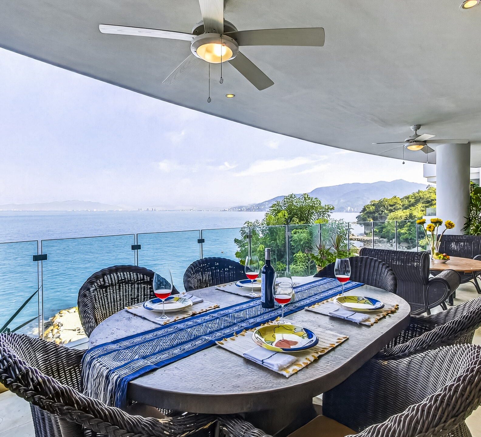 Covered terrace with wicker dining chairs and a table overlooking the coastline.
