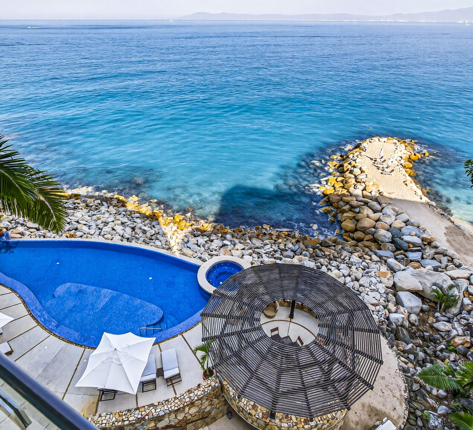 Aerial view of a swimming pool beside a rocky coastline and turquoise ocean.
