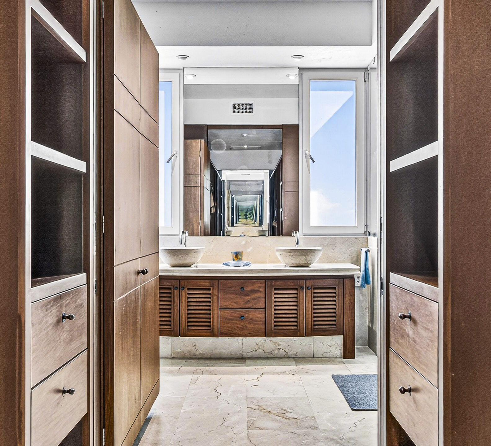 Walk-in closet with wood cabinetry leading to a spacious bathroom with double sinks.