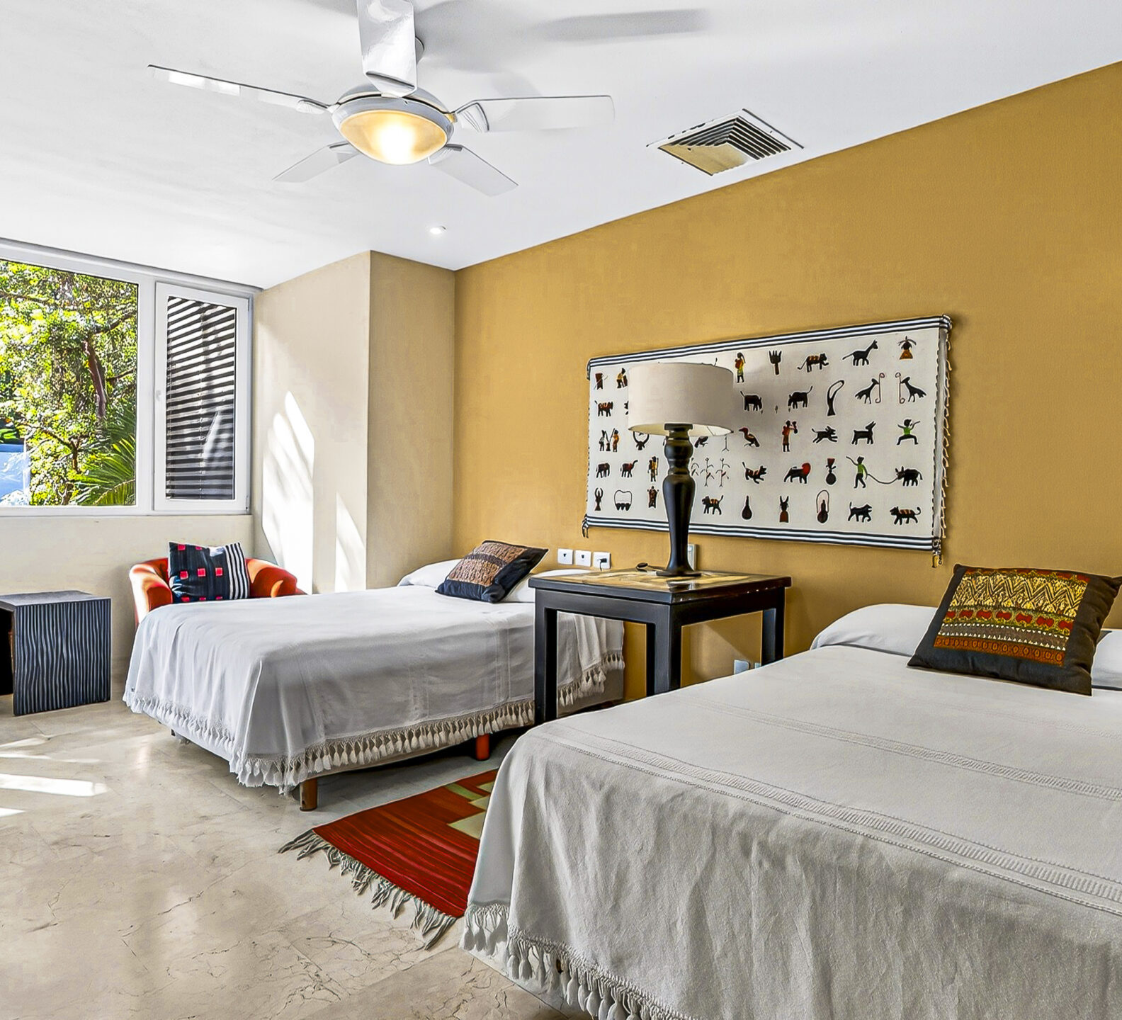 Guest bedroom with two beds, a desk, and large windows overlooking tropical greenery.