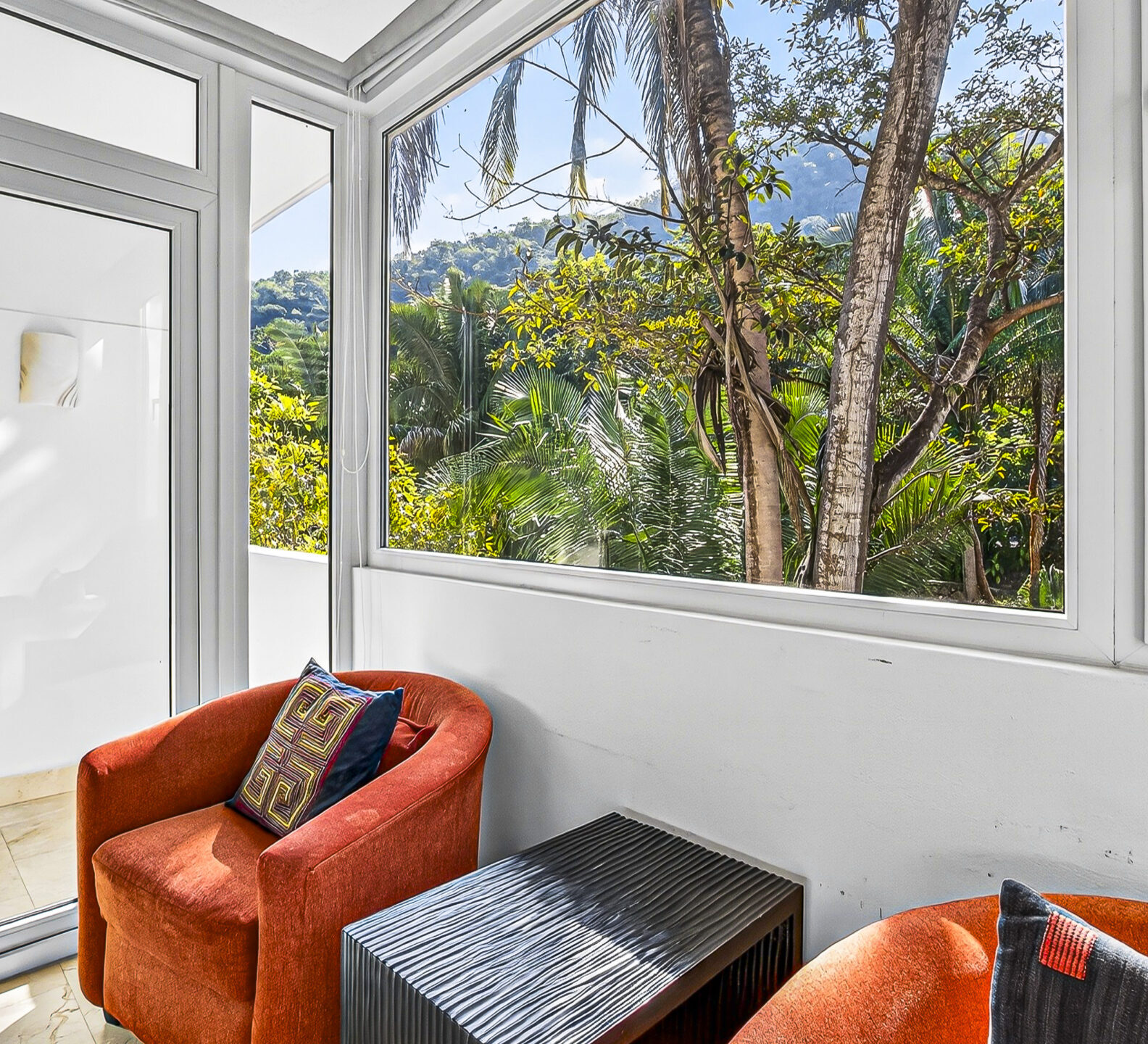 Sunlit sitting area with orange chairs and large windows overlooking tropical trees.