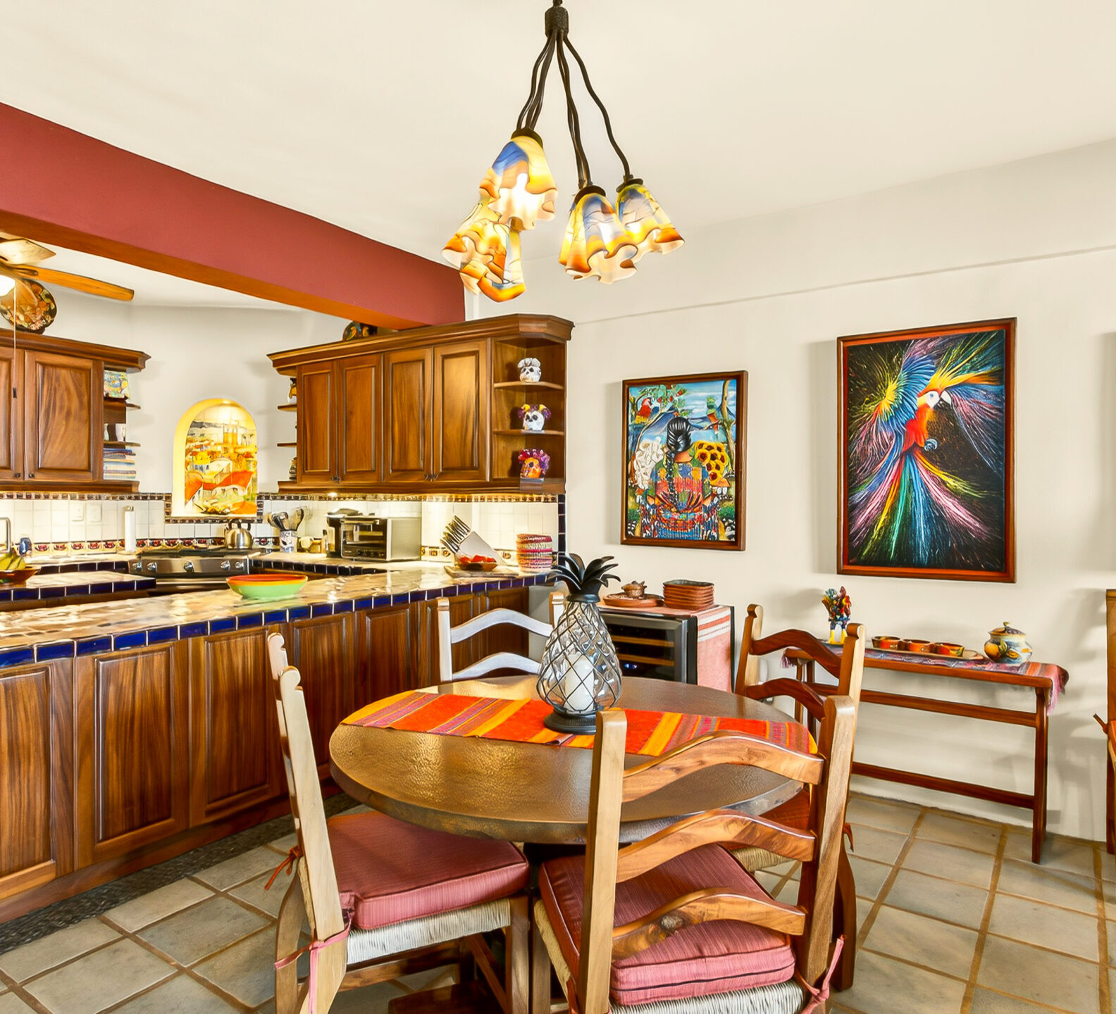 A round wooden dining table sits next to a kitchen with dark wood cabinetry and a tiled bar, accented by vibrant wall art.