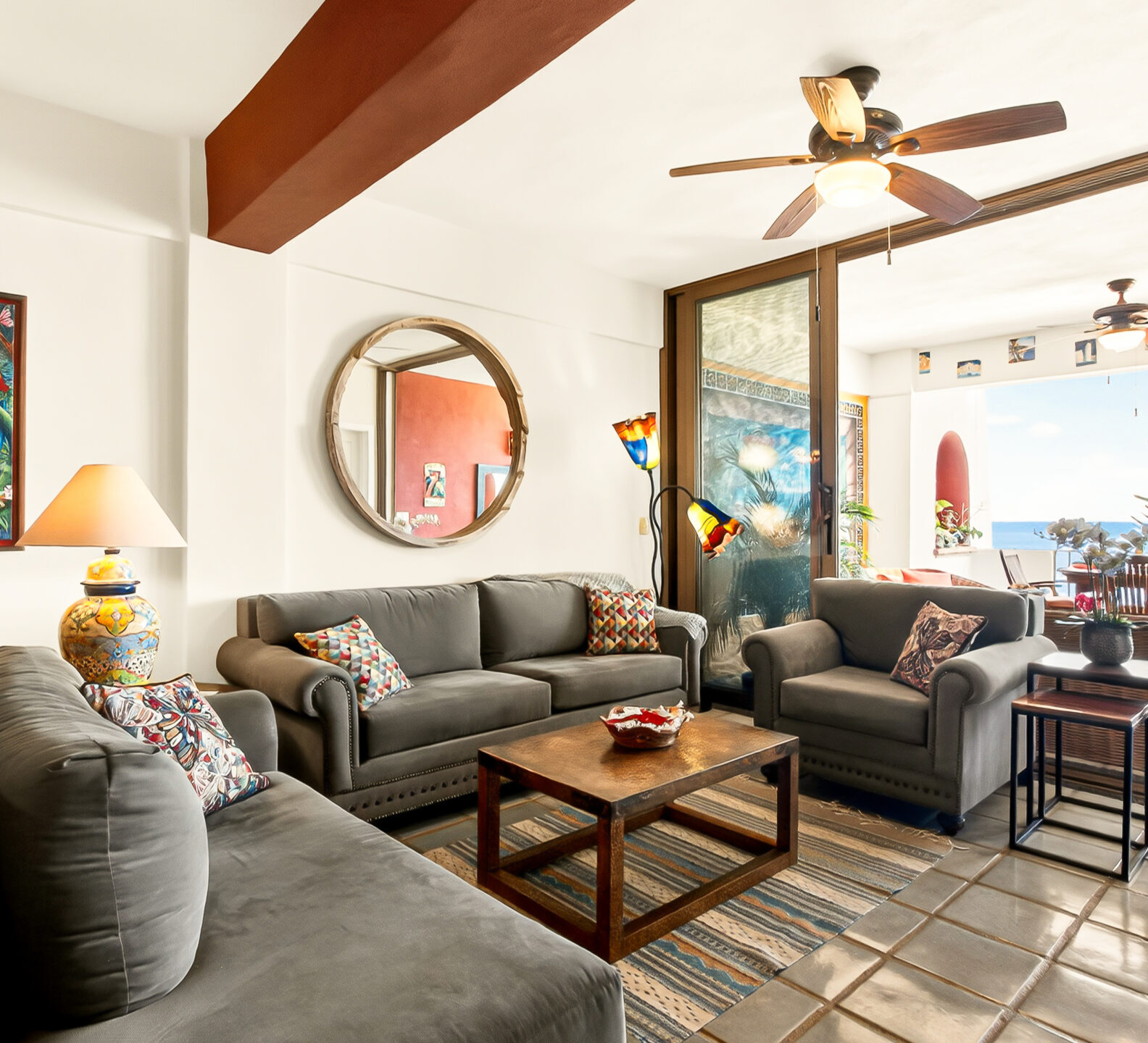 A cozy living room features grey sectional seating, a wooden coffee table, and large sliding doors opening to an outdoor patio with an ocean view.