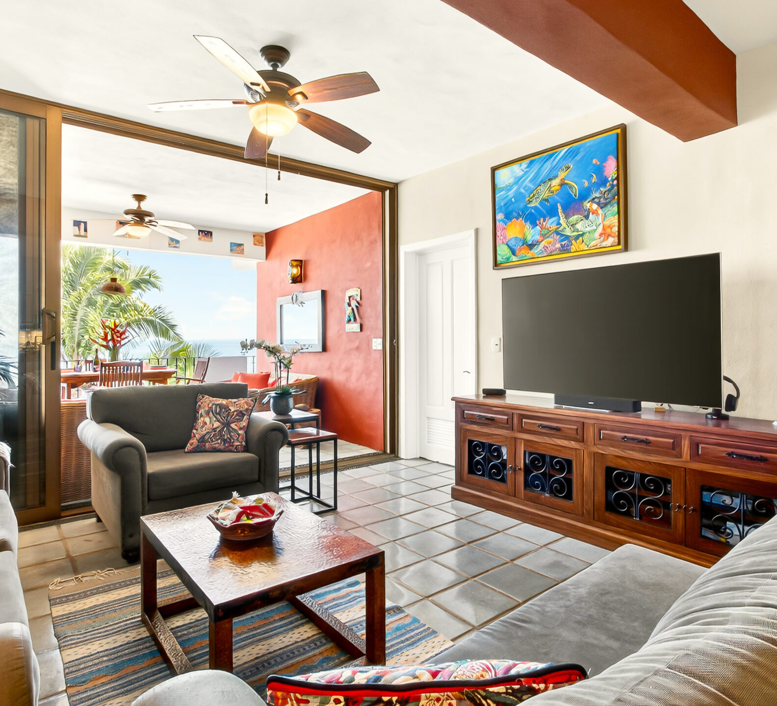 A living room with a large flat-screen television on a wooden media console, positioned next to open sliding doors revealing an outdoor terrace.