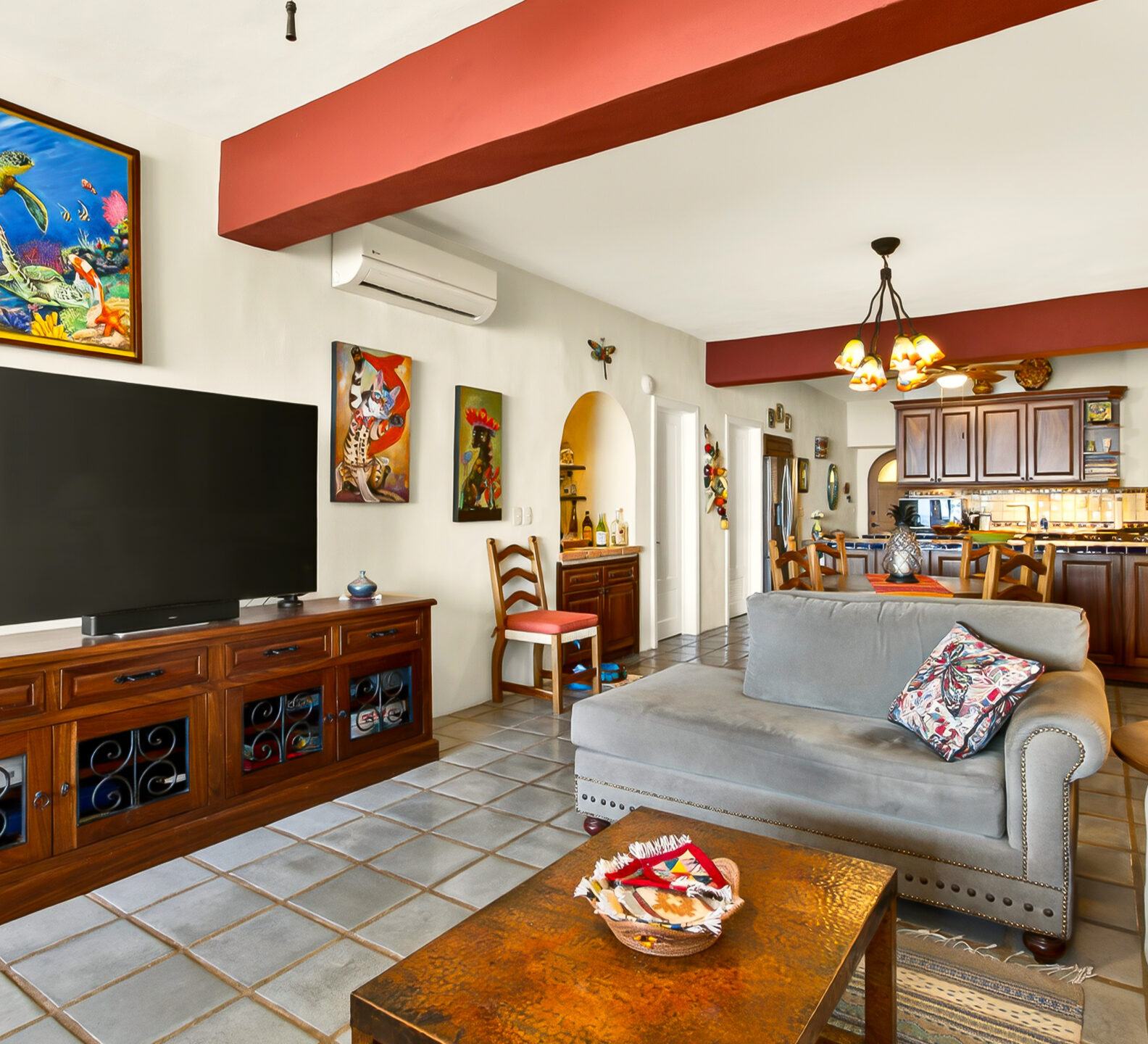 A view across the living room showing a large flat-screen television on a wooden media console, with the kitchen and dining area visible in the background.