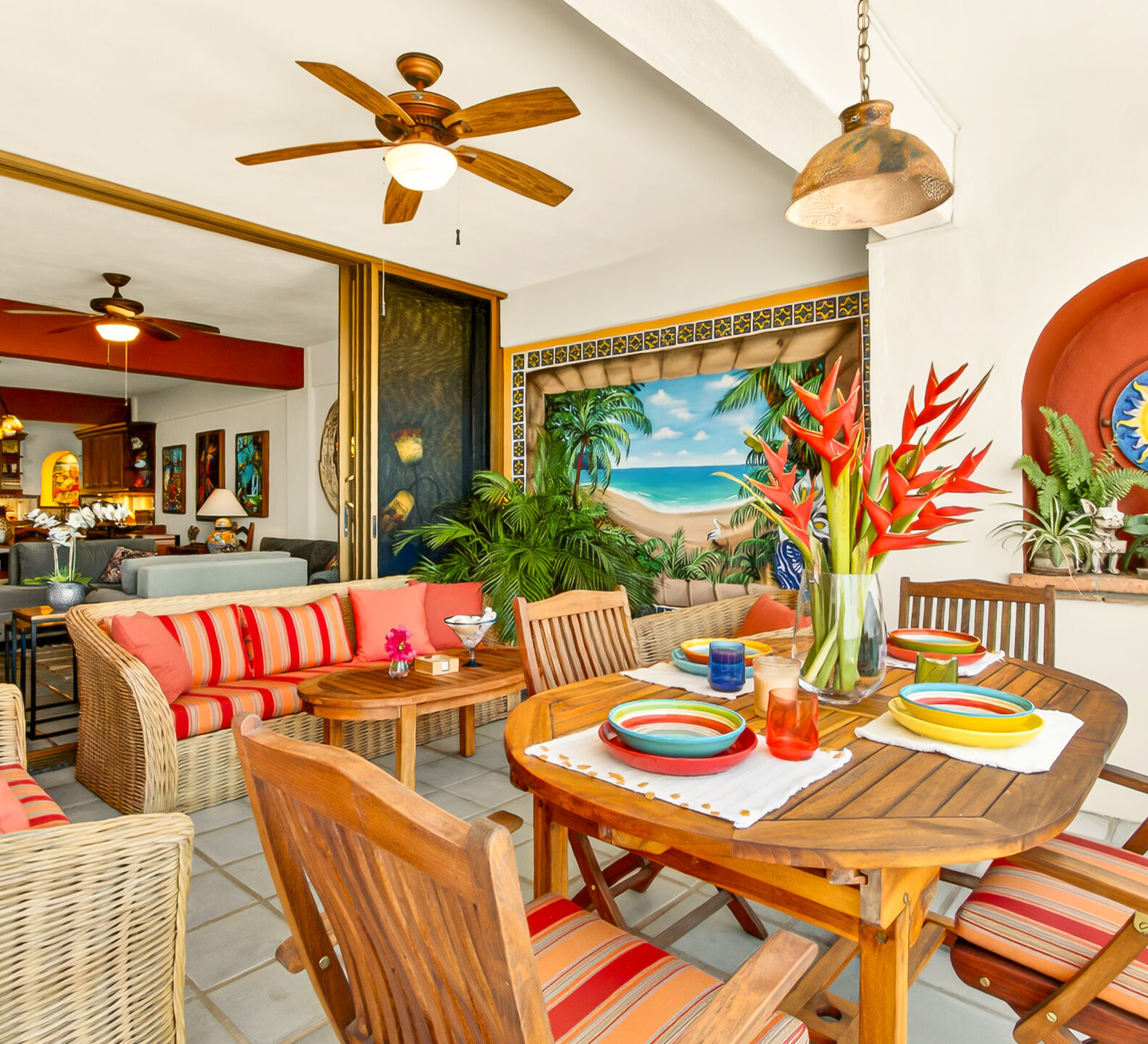 A covered outdoor patio featuring a round wooden dining table set for a meal and wicker lounge seating with vibrant orange striped cushions.