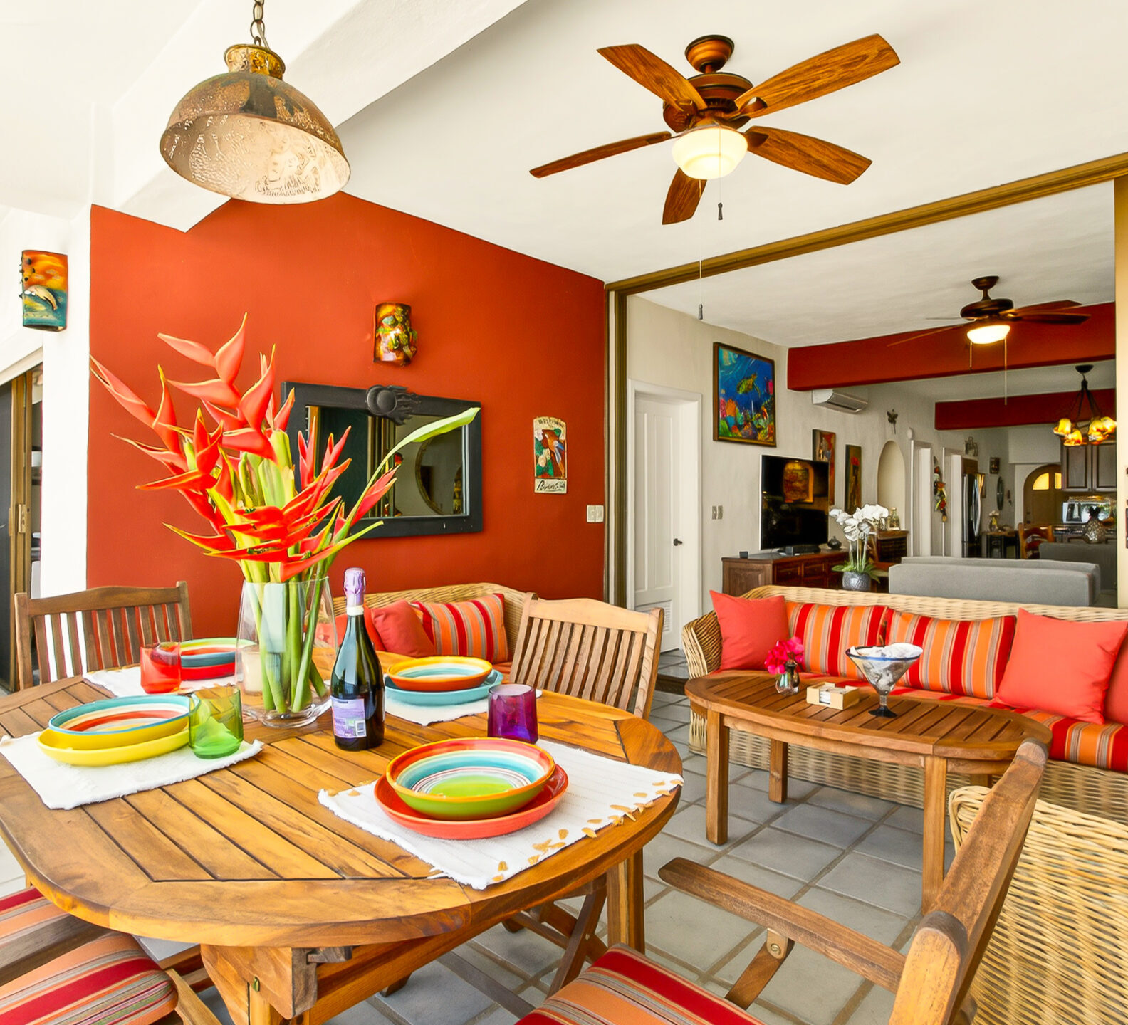 An outdoor dining area with a round wooden table, tropical floral centerpiece, and a view looking back into the open indoor living space.
