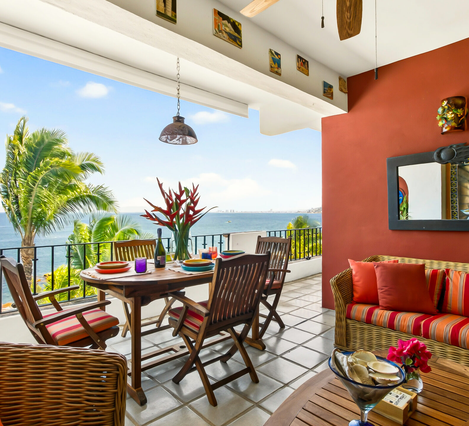 An outdoor terrace featuring a wooden dining table and chairs overlooking the beach and ocean, next to a lounge area with a red accent wall.