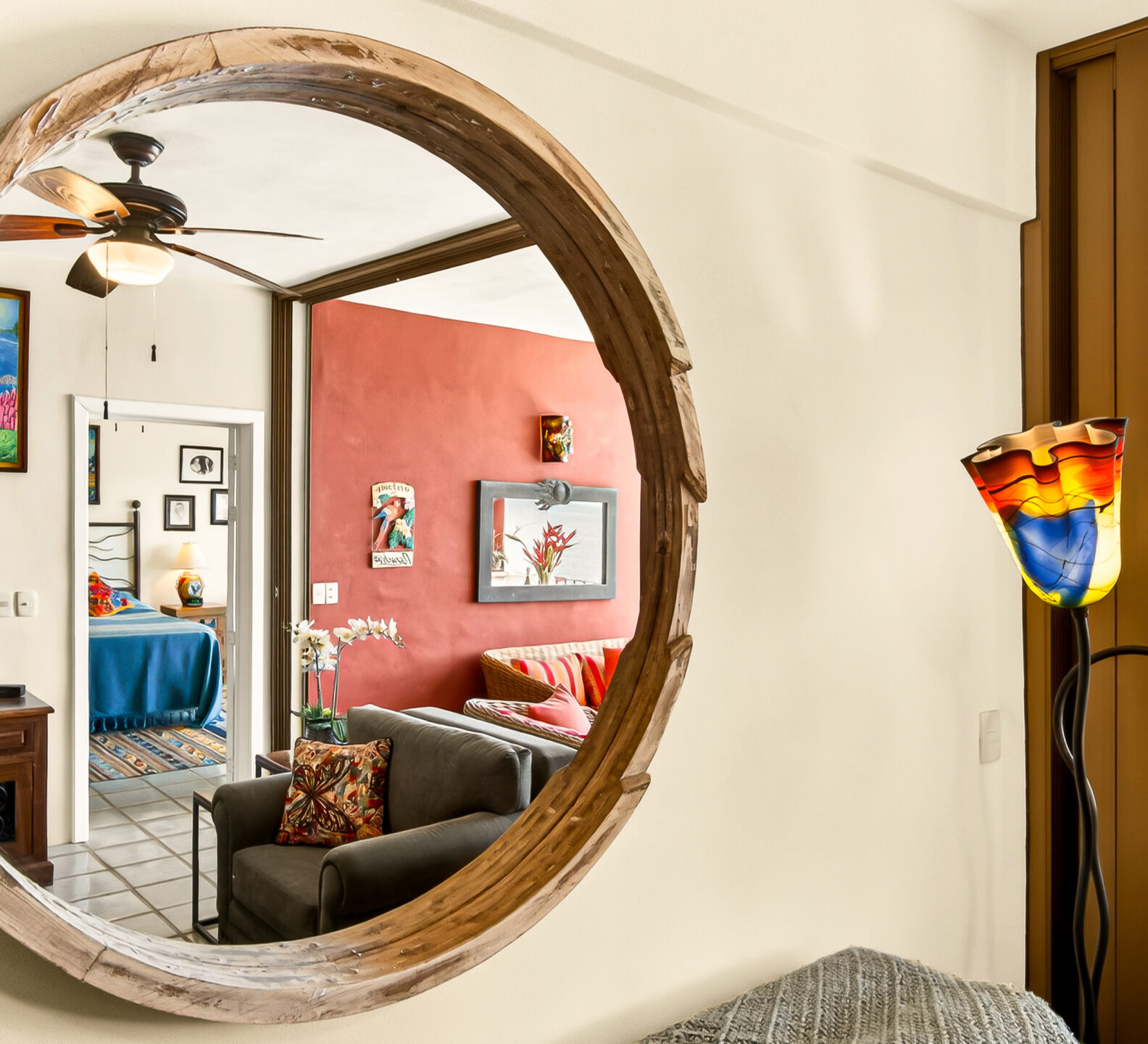 A large, round wooden-framed wall mirror reflecting an open-concept living area and an adjacent bedroom doorway.