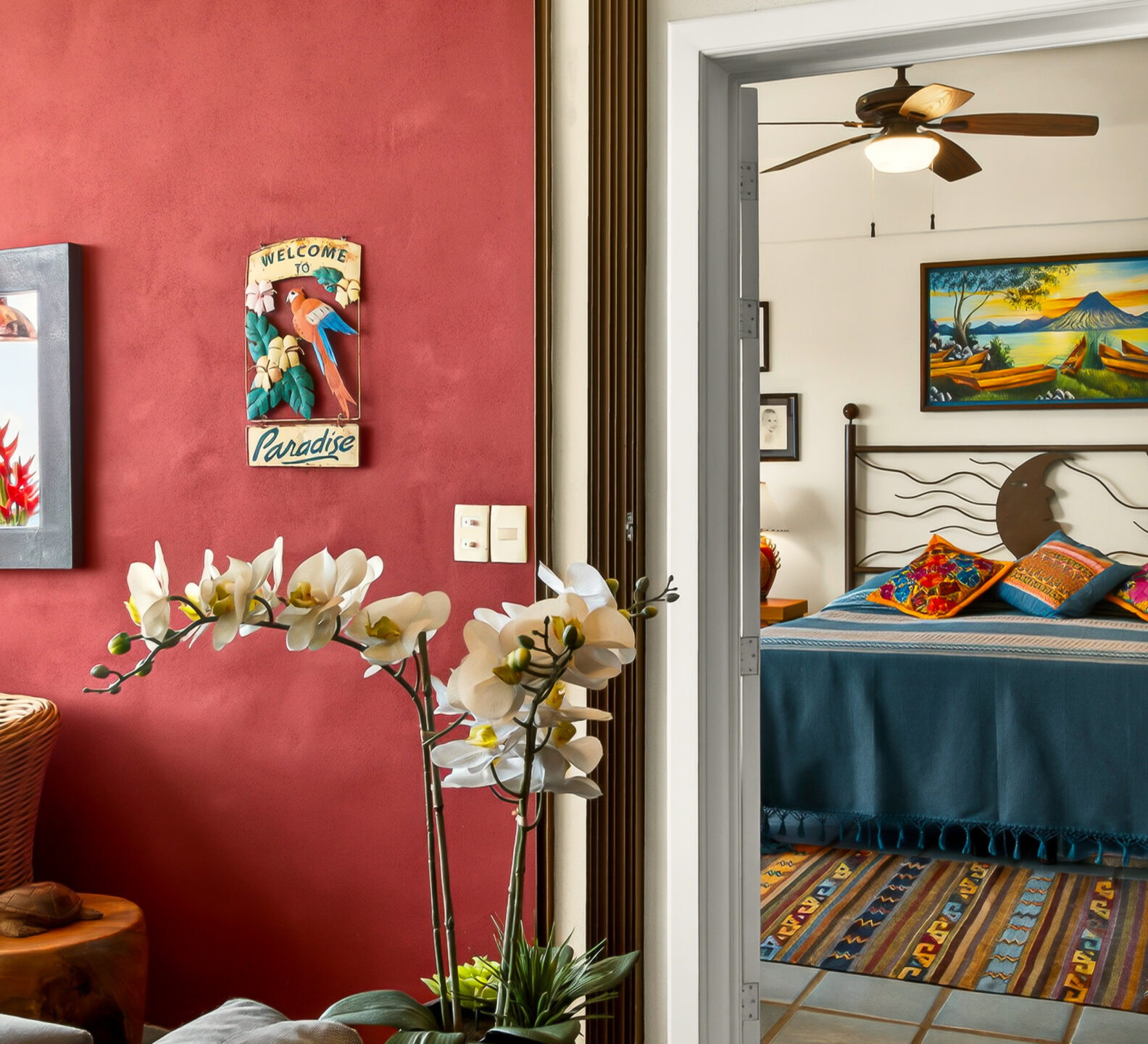 An open white doorway next to a red patio wall reveals a bedroom with a neatly made bed and a tropical painting hanging above it.
