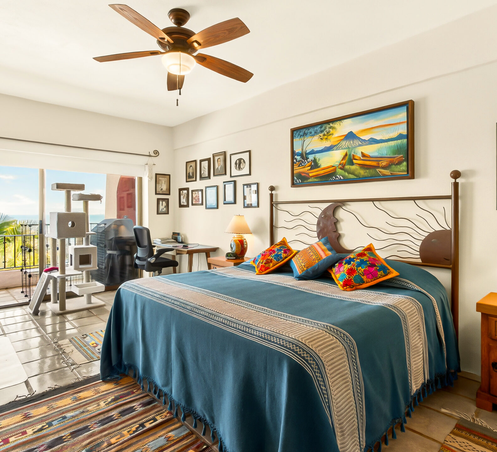 A spacious bedroom featuring a bed with a blue bedspread, a ceiling fan, and large sliding doors that open to a balcony overlooking the ocean.