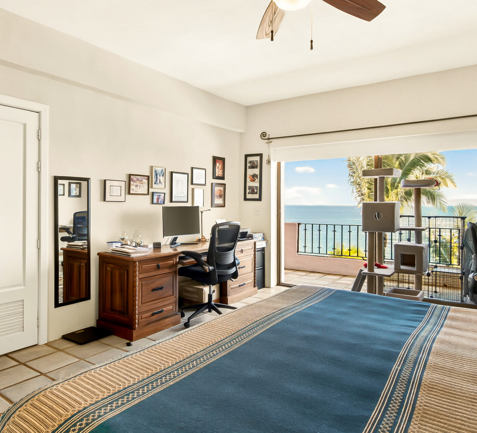 A bedroom view from the side of the bed, showing a wooden desk with a computer setup and large open sliding doors revealing an ocean view.