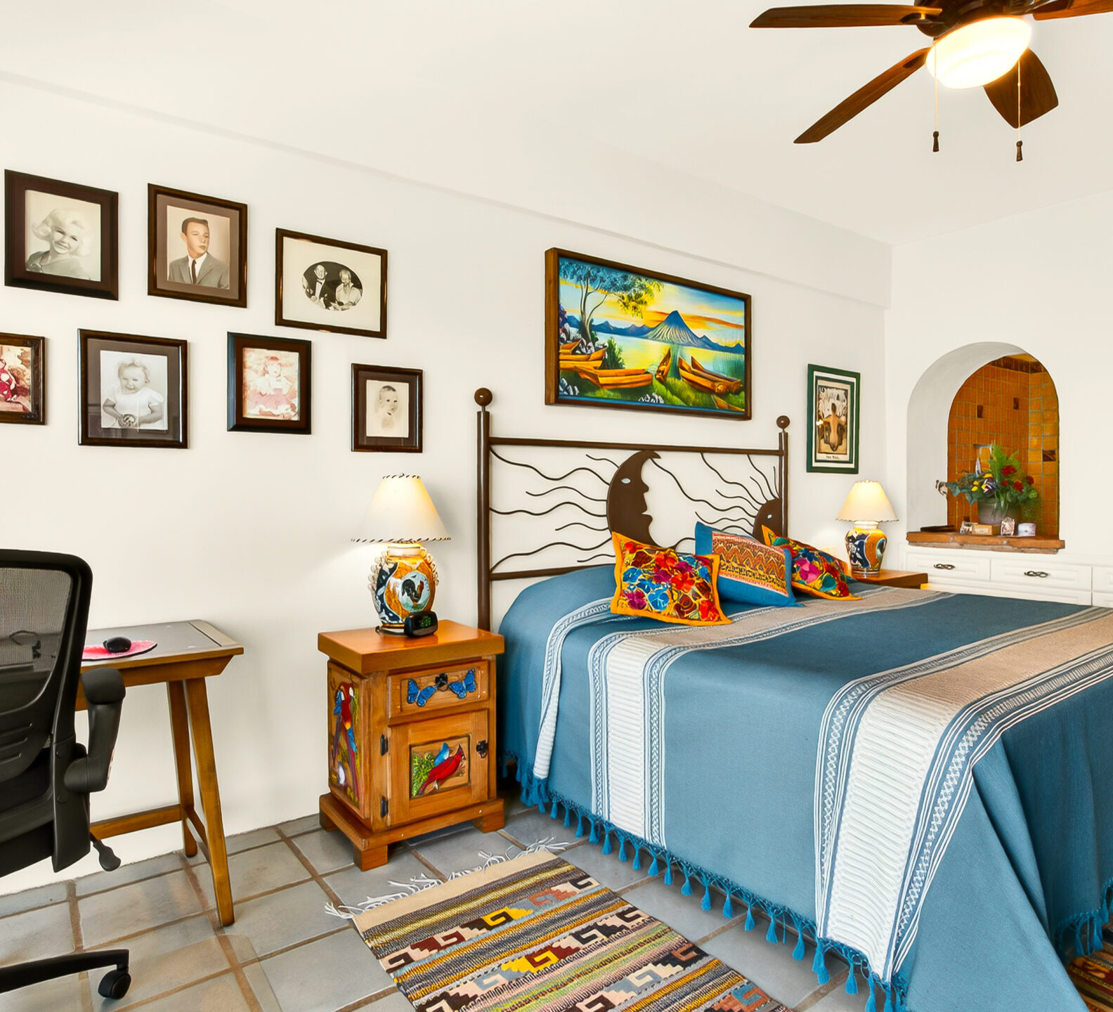 A bedroom featuring a bed with a blue and grey striped coverlet, a wall display of framed photos, and a black mesh office chair.