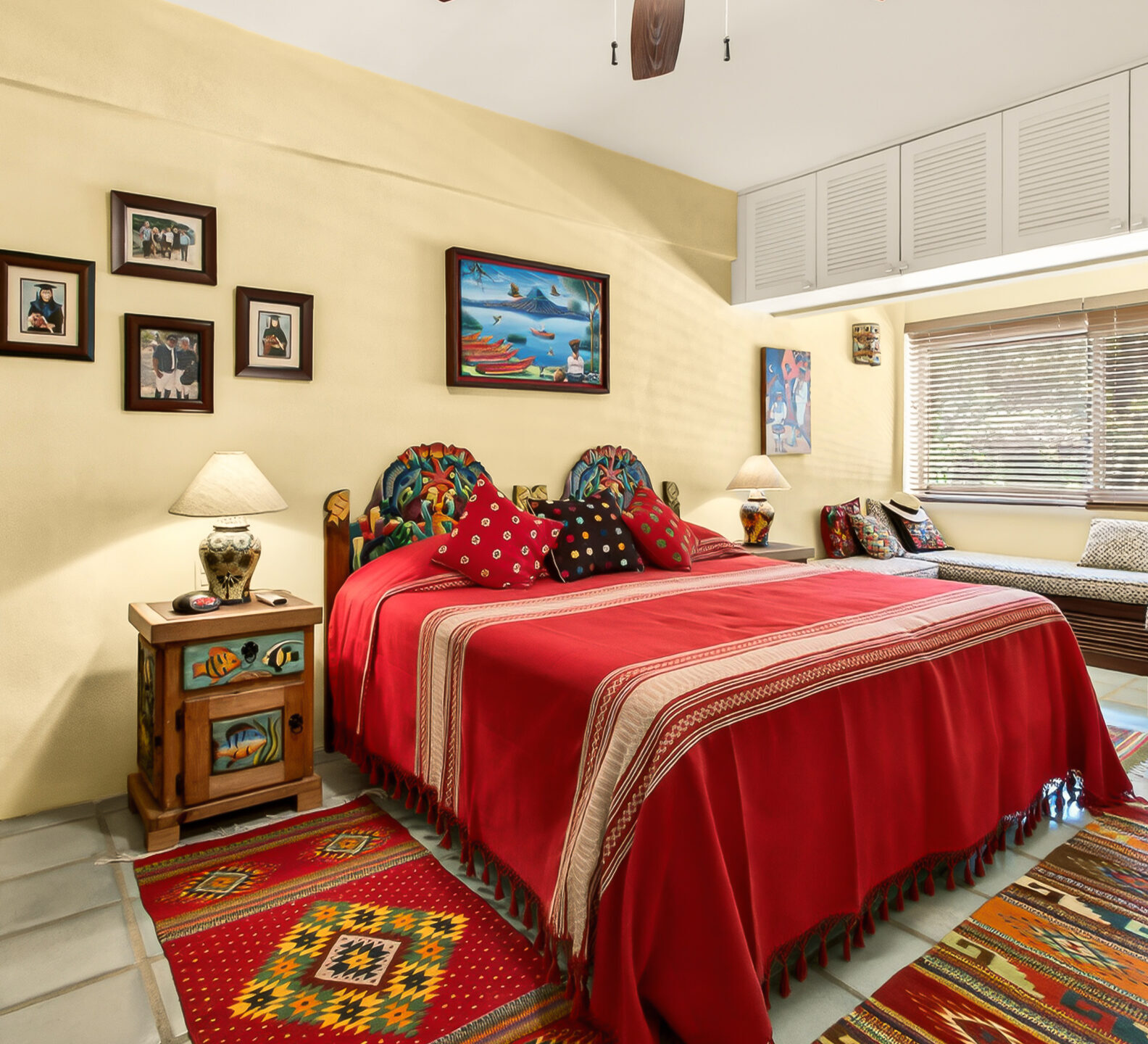 A bedroom featuring a bed with a vibrant red bedspread, warm beige walls decorated with artwork, and a window seat.