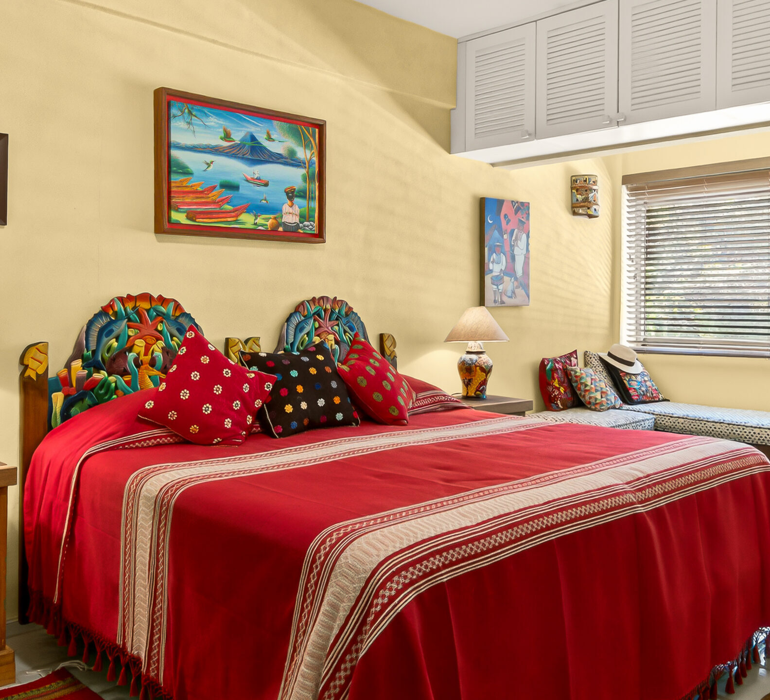 A close-up view of the bed with a red coverlet and an ornate headboard, positioned next to a window with a built-in bench.