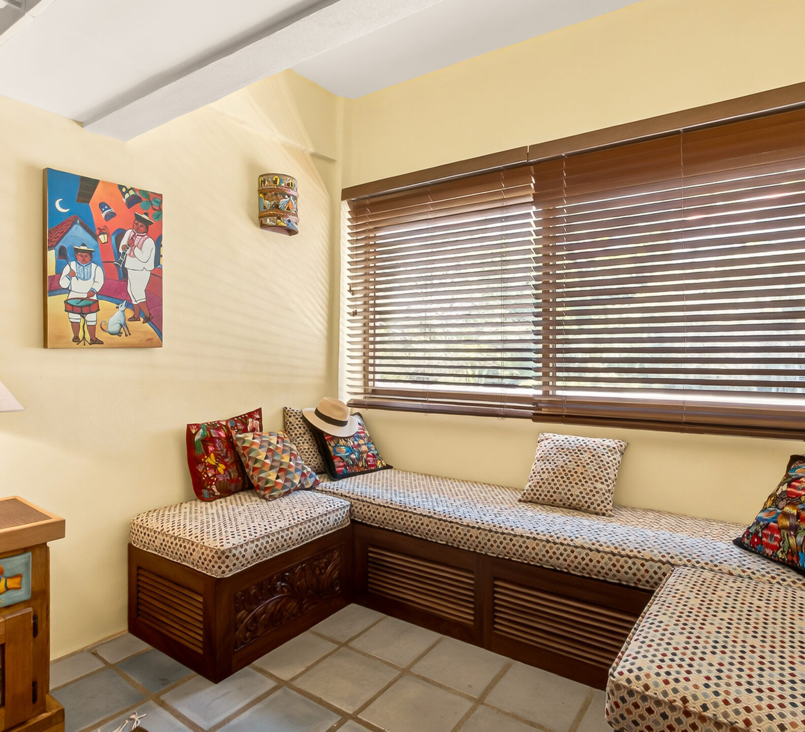 A cozy bedroom corner featuring a built-in wooden window bench with cushions and throw pillows beneath closed blinds.