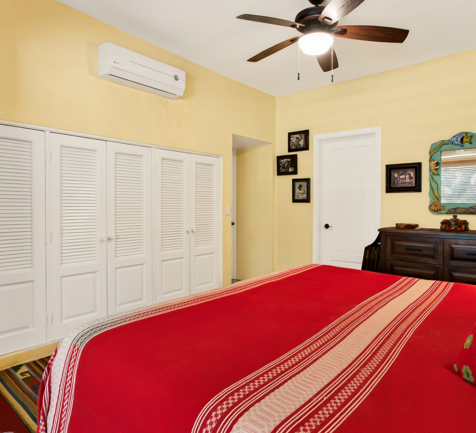 A bedroom view from the side of a red-covered bed, showing a long wall of white louvered closet doors and a ceiling fan.
