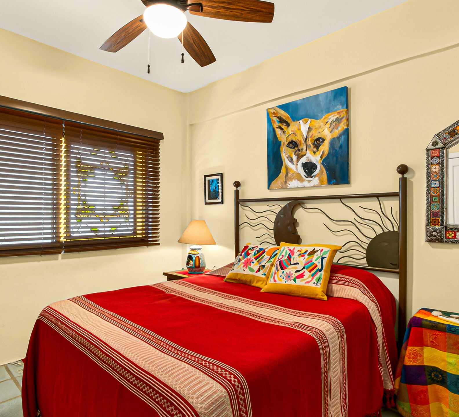 A bedroom featuring a bed with a red-and-orange striped coverlet, closed wooden blinds, and a painting of a dog above the headboard.