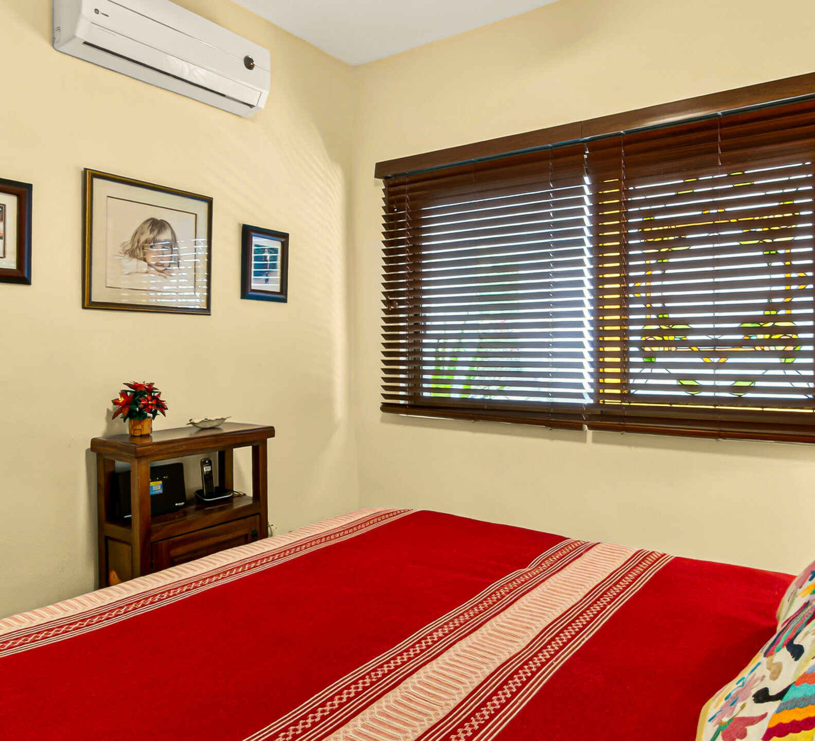 A close-up view over a red bed toward a window with dark wood blinds, an air conditioner, and framed art on a beige wall.