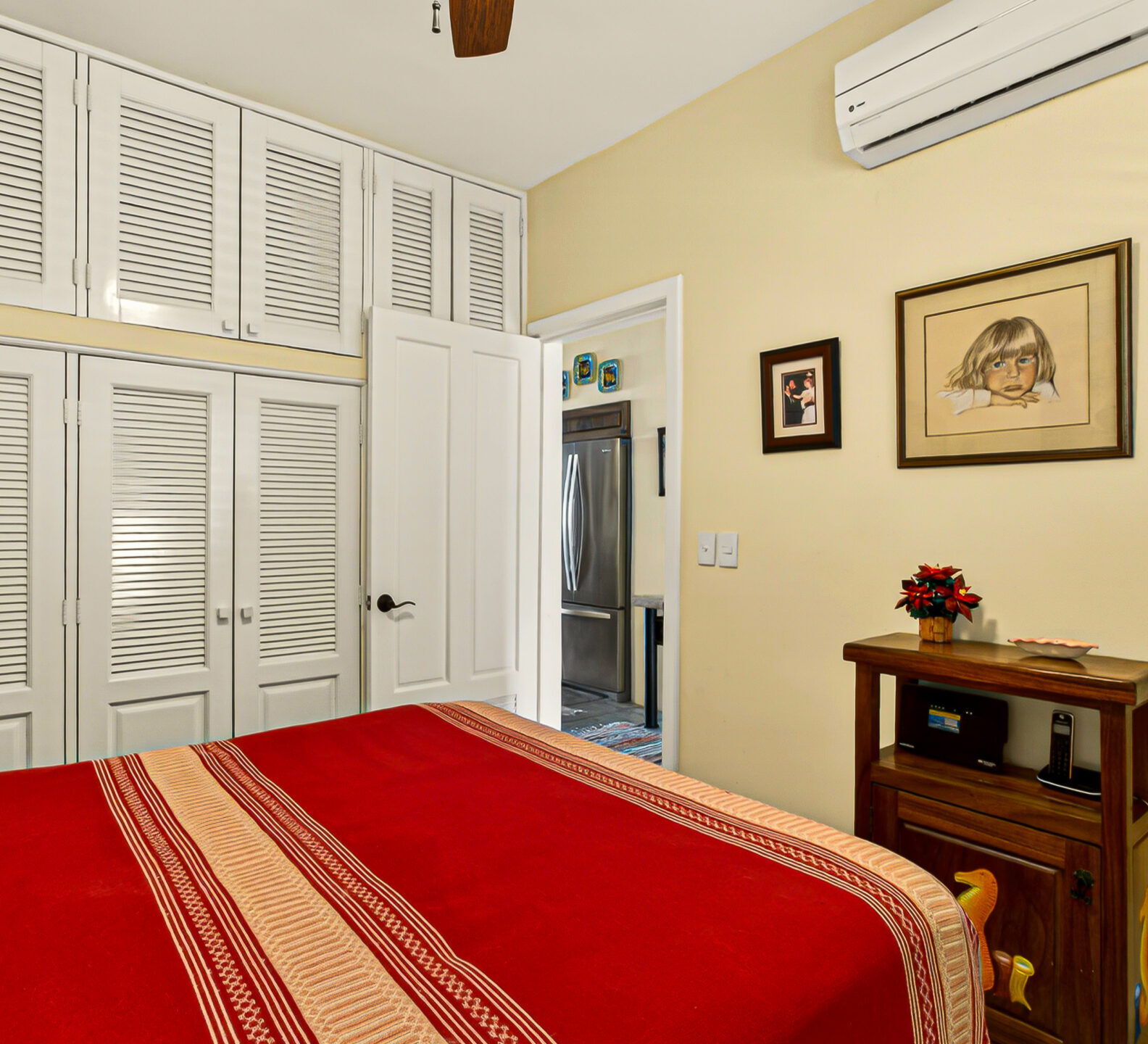 A bedroom view showing a red bed facing a wooden accent table, with a large wall of white louvered closet doors extending to the ceiling.