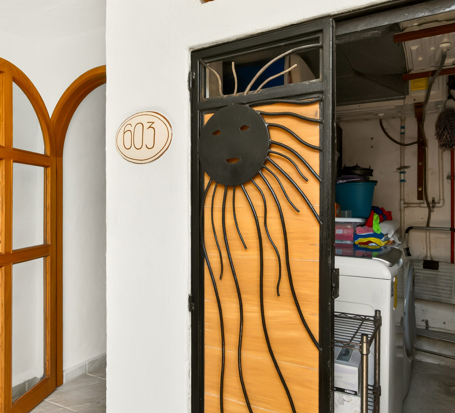 An exterior entryway featuring an arched wooden glass door and an adjacent laundry or utility closet with a decorative sun-motif metal gate.