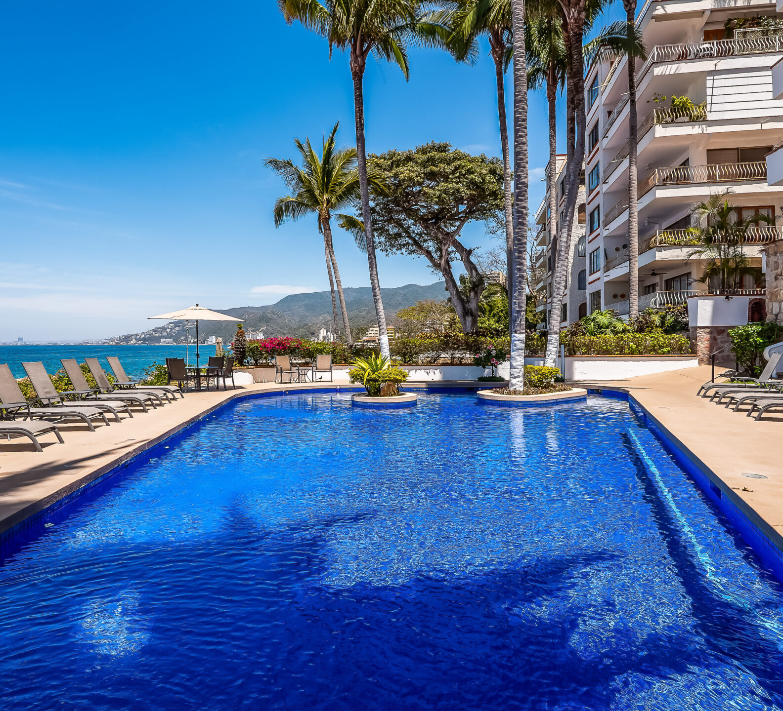 A sparkling blue swimming pool lined with lounge chairs sits adjacent to a multi-story beachside resort building under a clear sky.