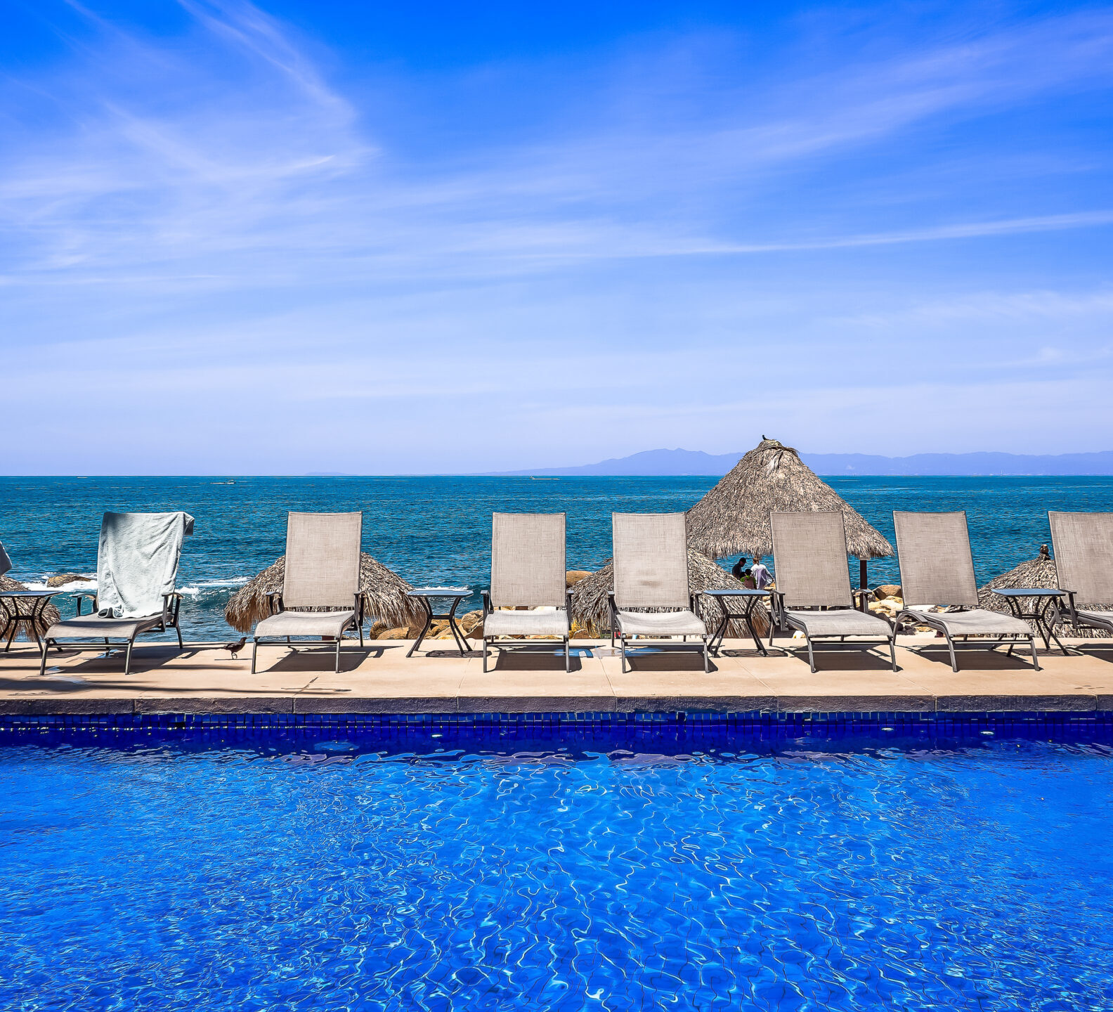 A row of white lounge chairs sits on a sun deck overlooking a blue swimming pool and the expansive ocean beyond.