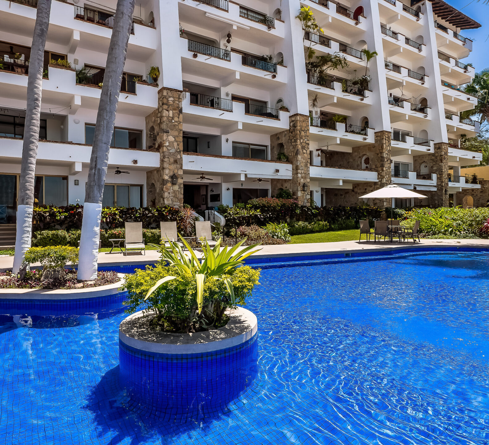 A curved swimming pool featuring a small central palm tree planter reflects the multi-story facade of a modern beachfront condominium.