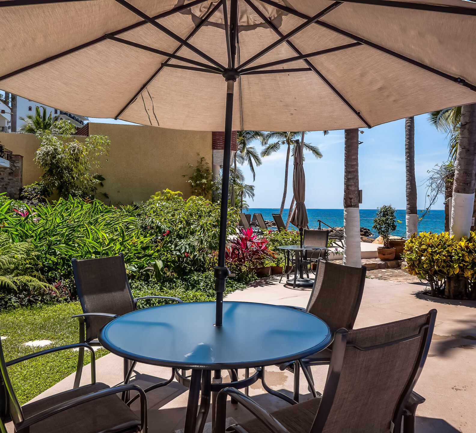 An outdoor patio table with four chairs sits shaded under a large umbrella next to a garden walkway and the pool.