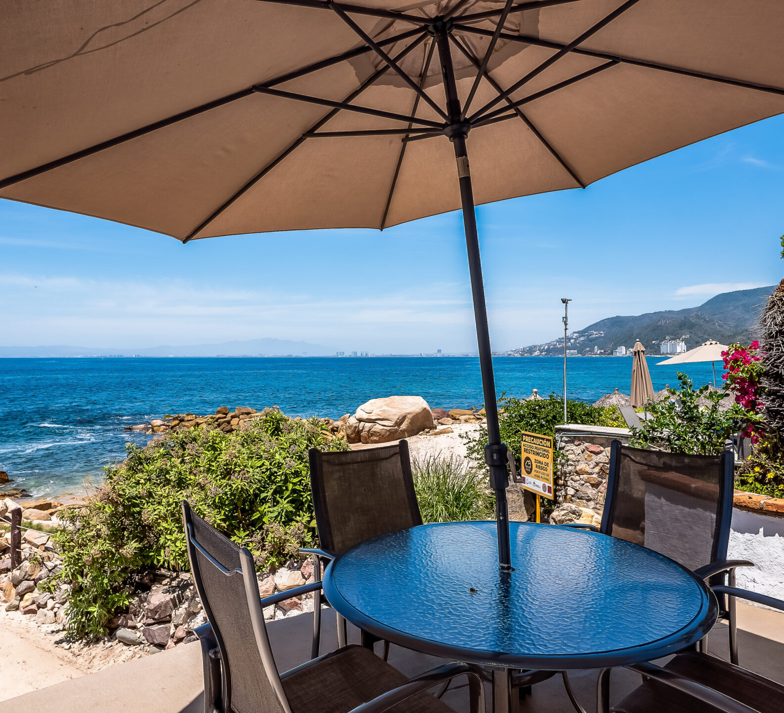 A round outdoor dining table under a large patio umbrella offers panoramic views of the rocky coastline and blue ocean.