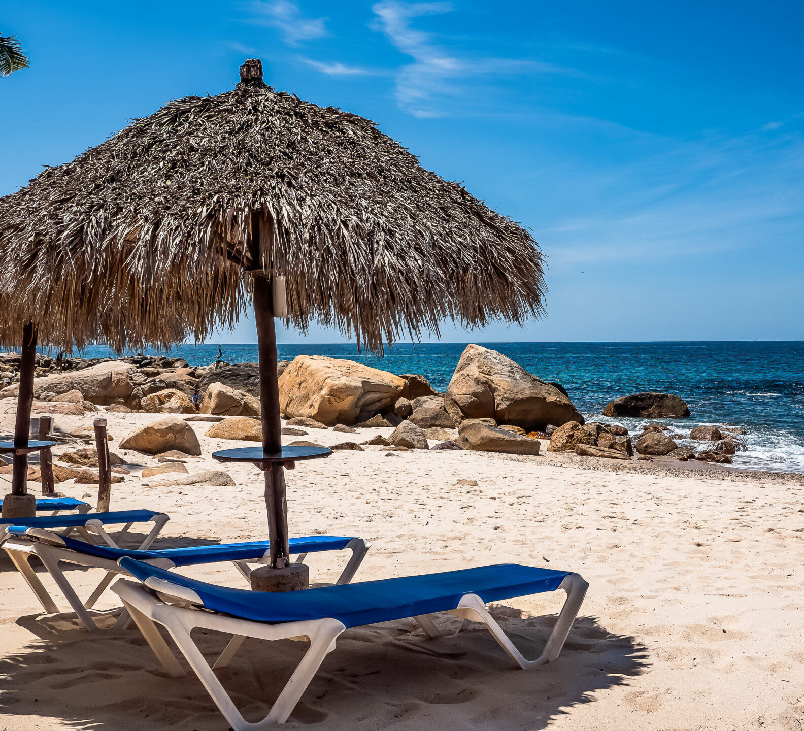 Blue lounge chairs sit on a sandy beach under thatched palapa umbrellas overlooking the ocean waves and a rocky shoreline.