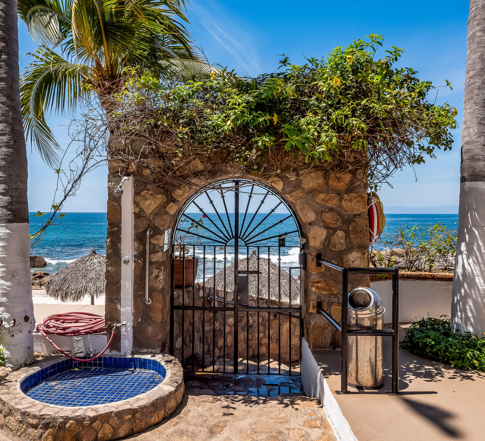 An ornate stone archway with a black metal gate leads directly out to a sandy beach lined with palm trees.