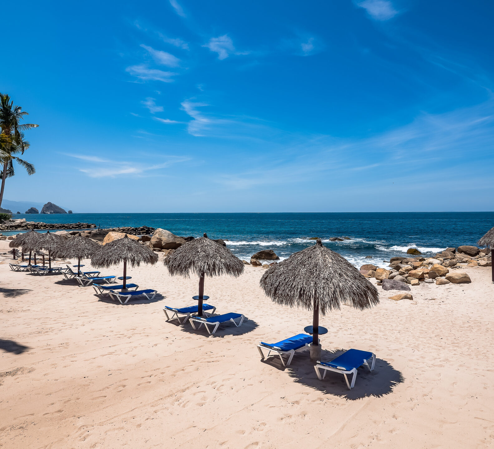 A wide, elevated view of a sandy beach resort area dotted with thatched palapa umbrellas and lounge chairs next to the ocean.