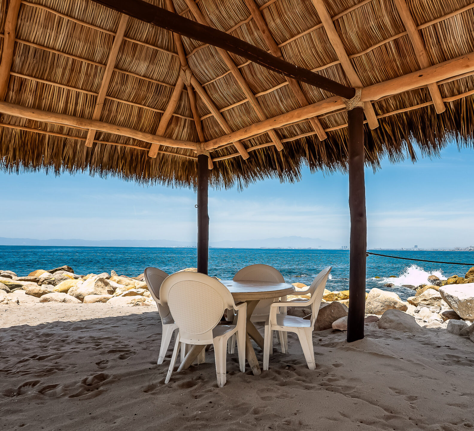 A shaded seating area under a large thatched palapa roof features white plastic chairs and a table right along the rocky ocean shoreline.