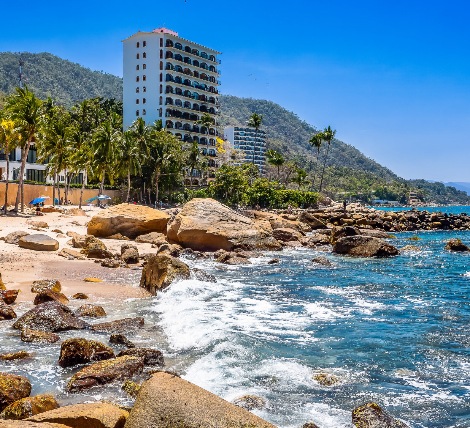Ocean waves crash against a rocky shoreline next to a sandy beach with palm trees and a tall white resort building in the distance.