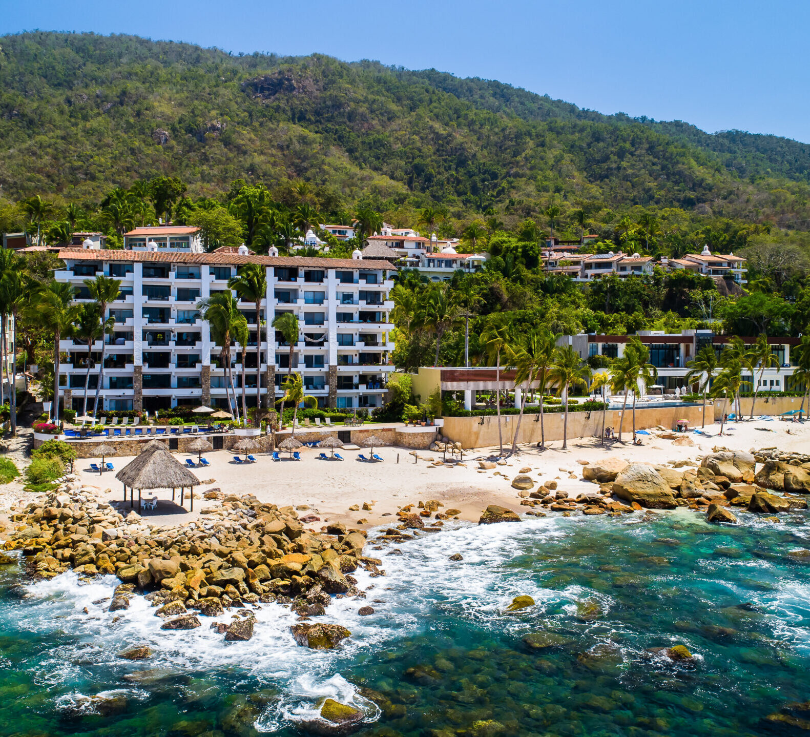 An aerial view showcases a multi-story beachfront condominium complex nestled along a rocky cove with lush green mountains in the background.