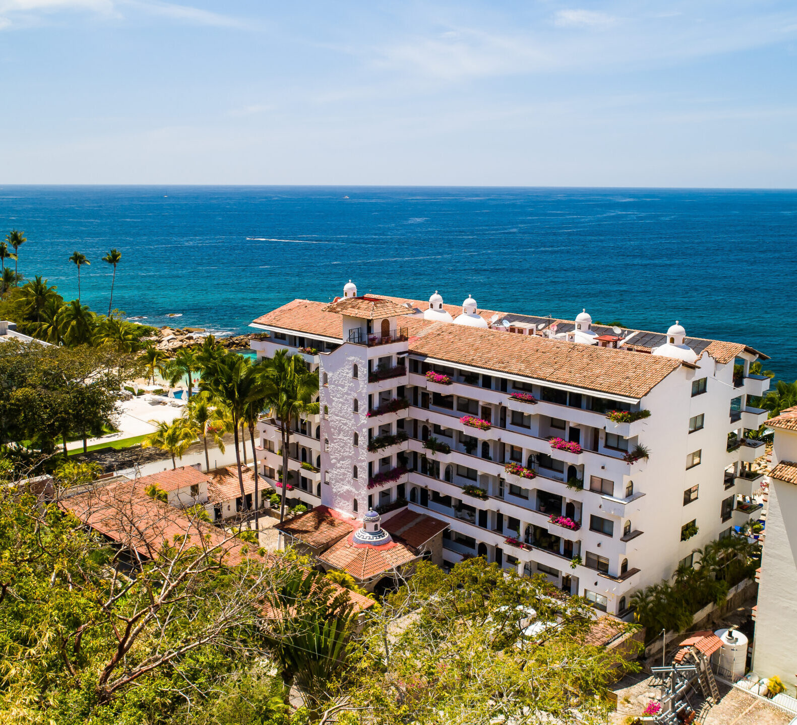 A high-angle view looks down at a multi-story resort complex with a terracotta tiled roof sitting right along the deep blue ocean coastline.