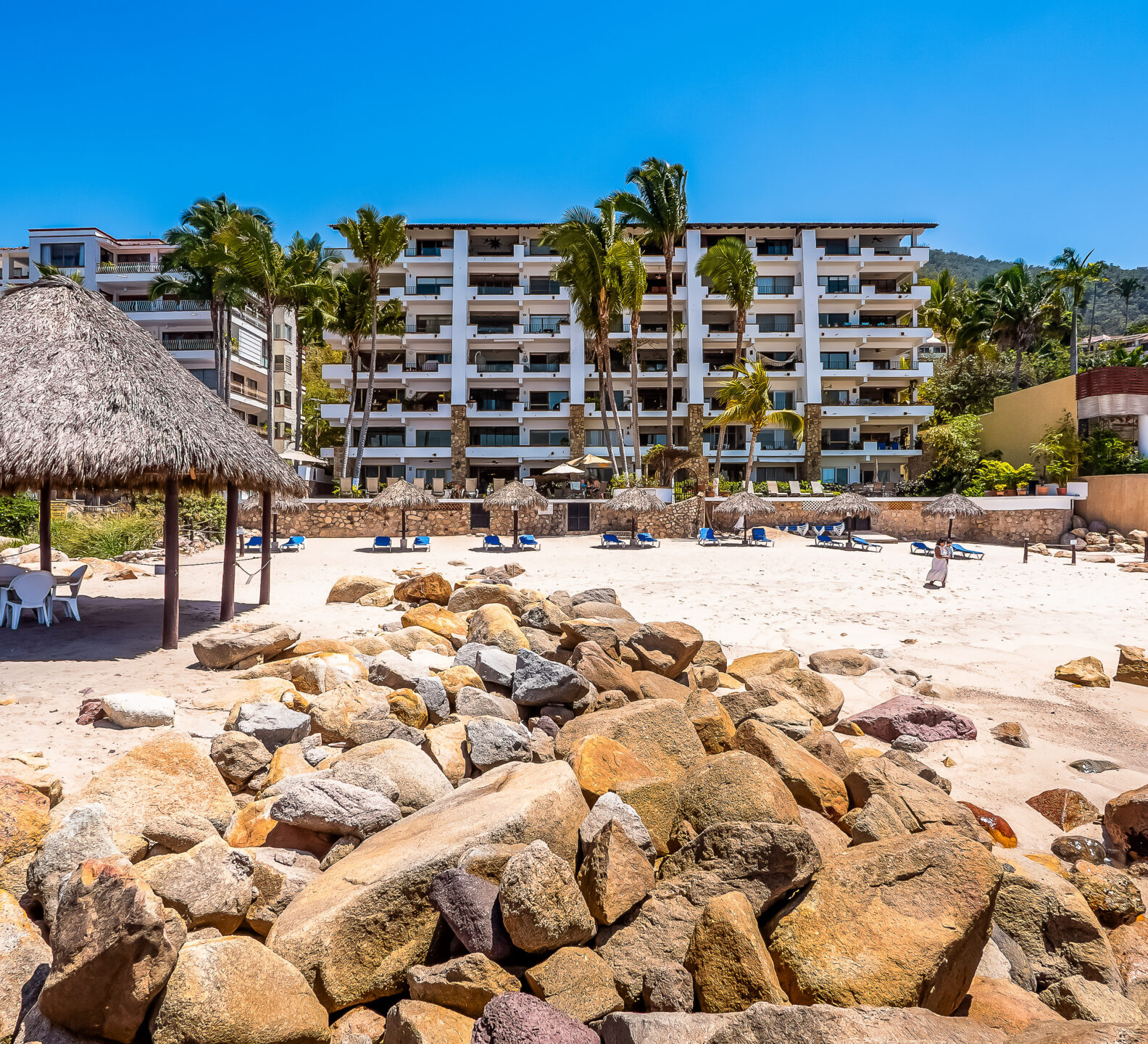 A large thatched palapa structure sits on a sandy beach behind a rocky shoreline, with a white resort building in the background.