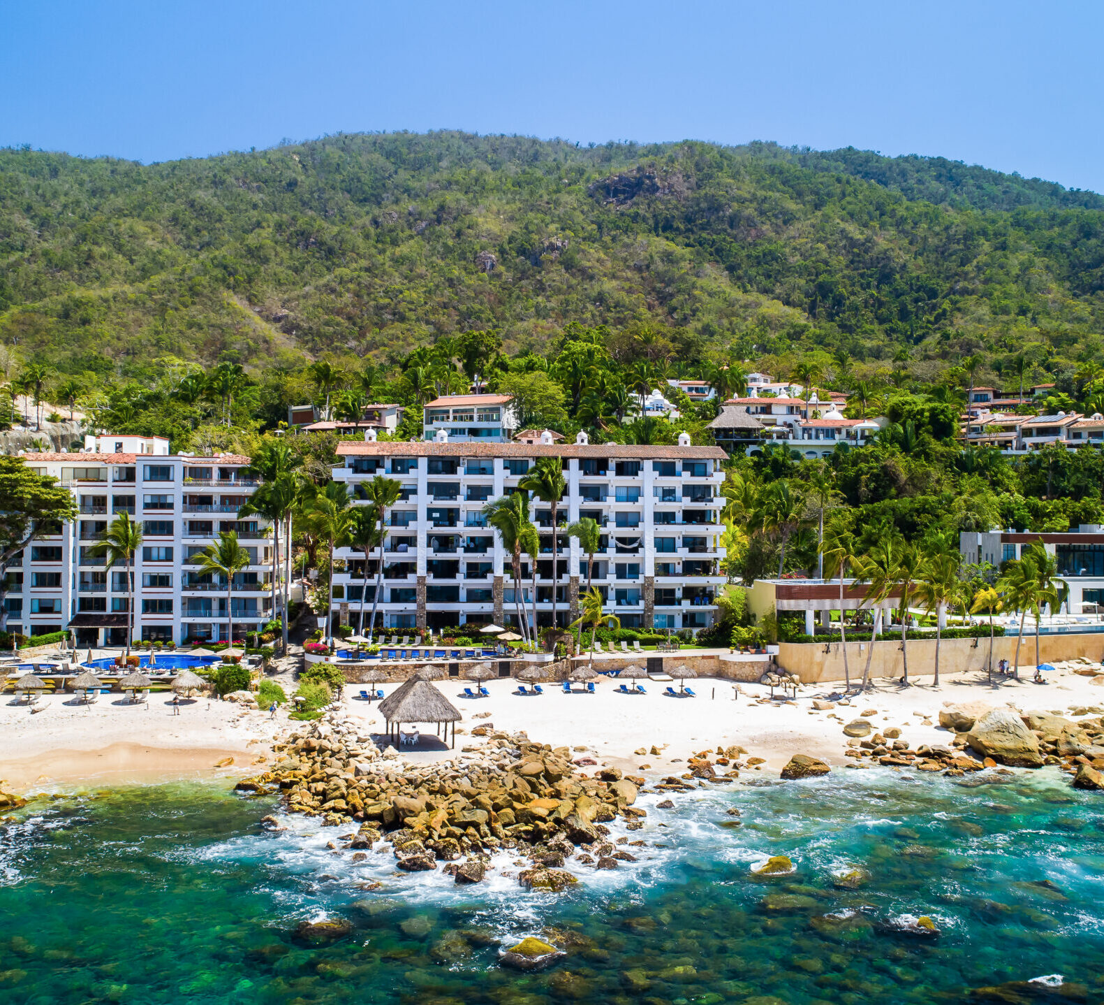 An aerial coastal view reveals a multi-story beachfront condominium resort nestled between turquoise ocean waters and lush green mountains.