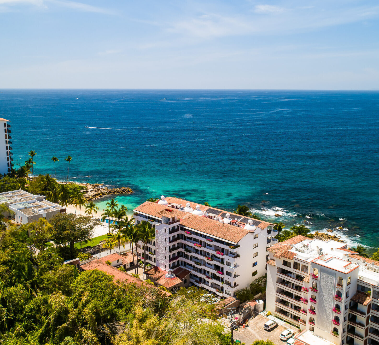 A high-angle panoramic view shows multiple resort buildings situated along a vibrant blue ocean coastline framed by lush green trees.