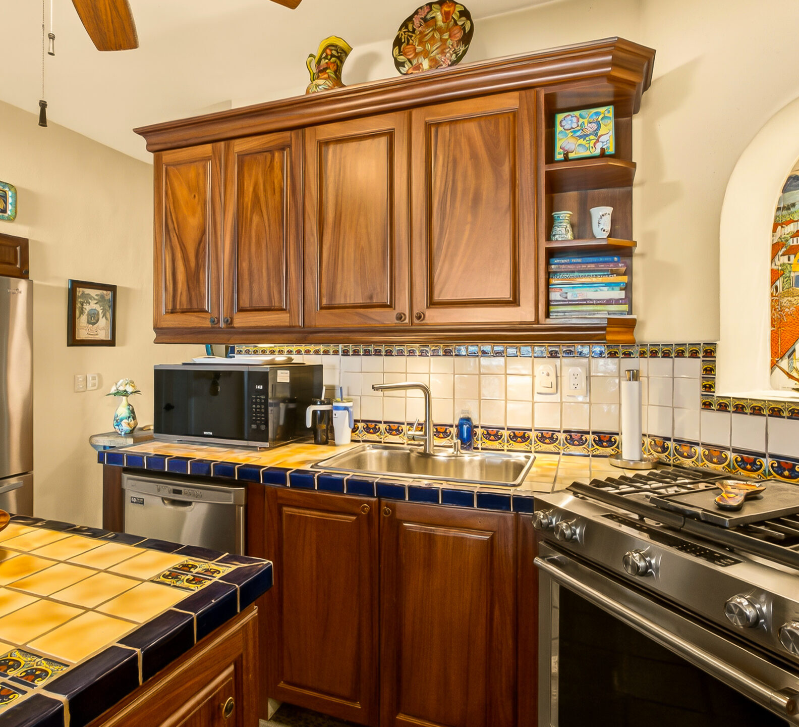 Warm, traditional kitchen with wooden cabinets, tiled countertops, and a colorful arched window above the sink.