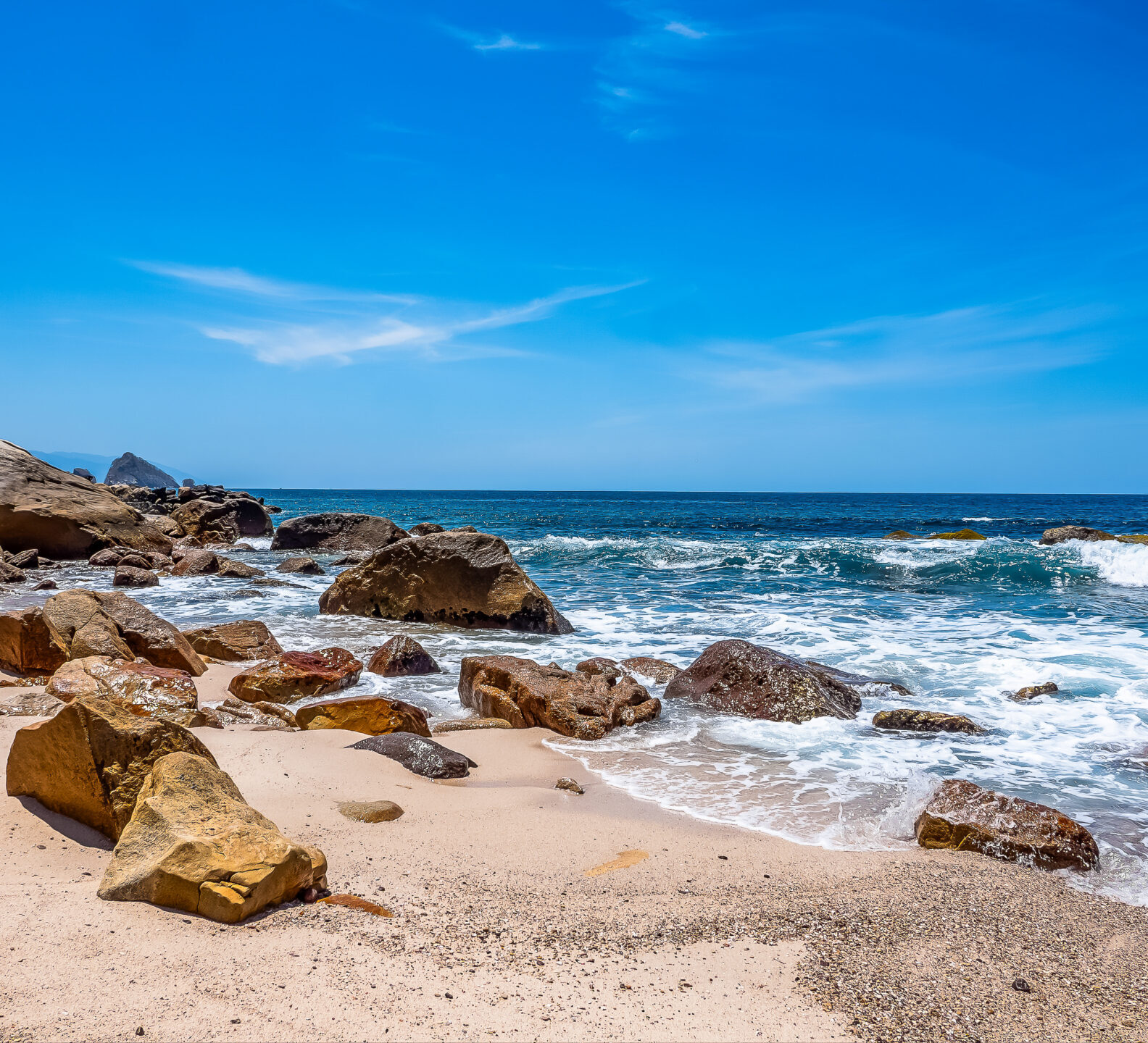 Ocean waves crash onto a sandy beach dotted with large tan boulders under a vibrant blue sky.