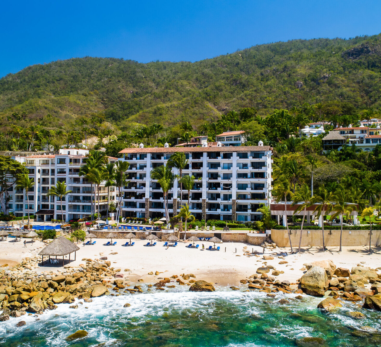 A wide coastal shot showcases a white multi-story resort building built against a steep, lush green mountain backdrop.