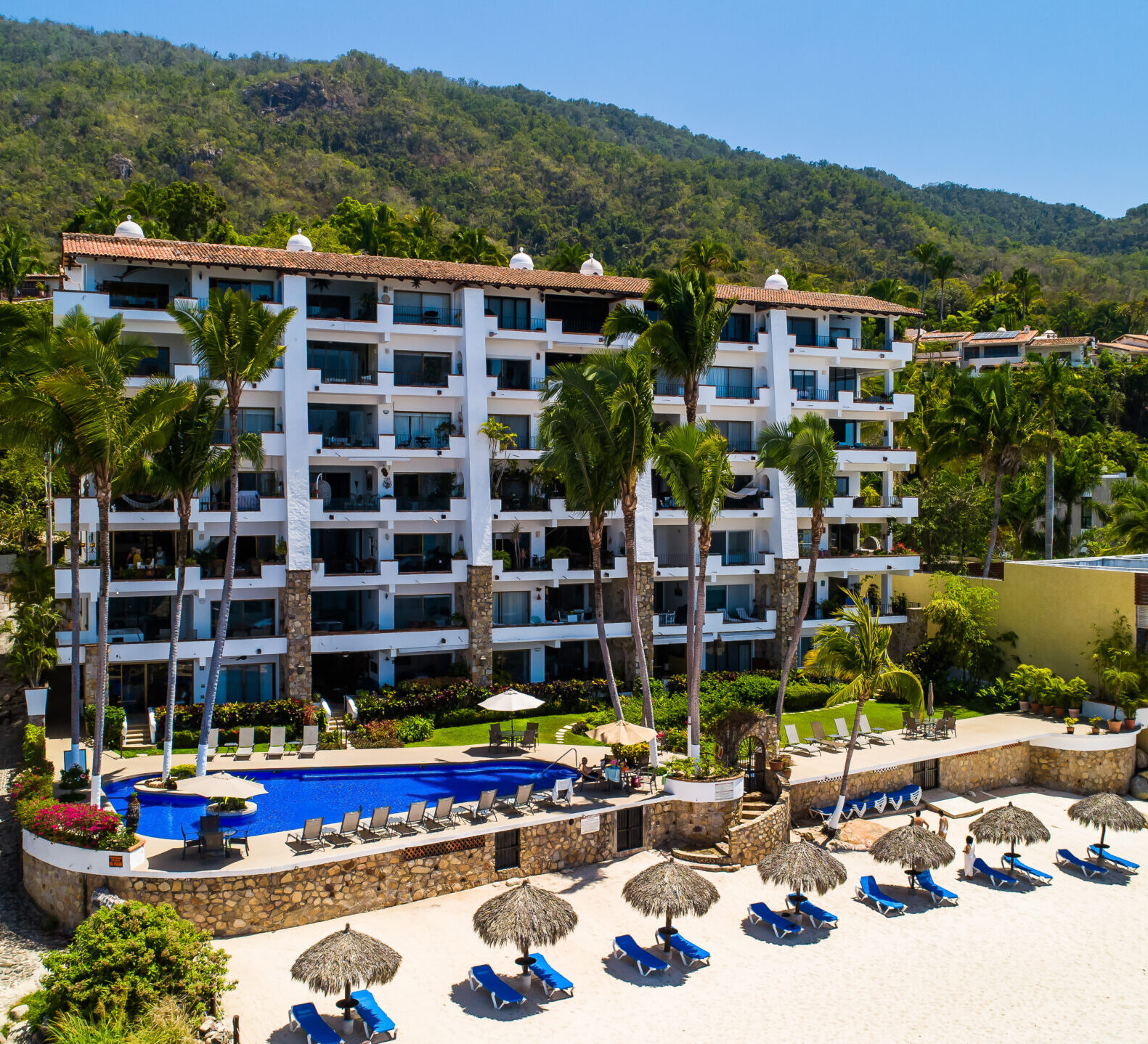 An elevated view shows a white multi-story resort with a blue swimming pool and a sandy beach lined with palapa umbrellas.