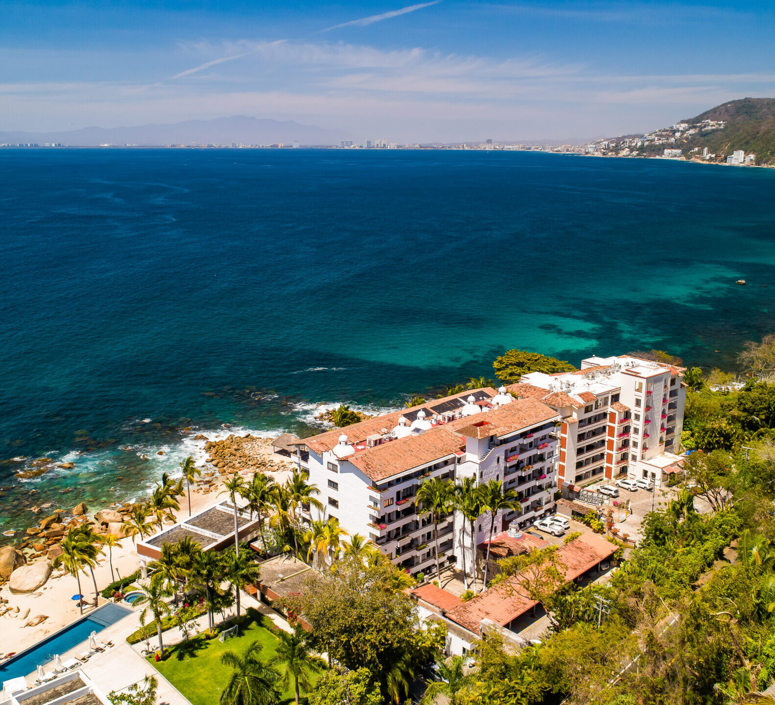 A high-angle panoramic shot overlooks a coastal resort building nestled in greenery next to a vast, turquoise ocean bay.