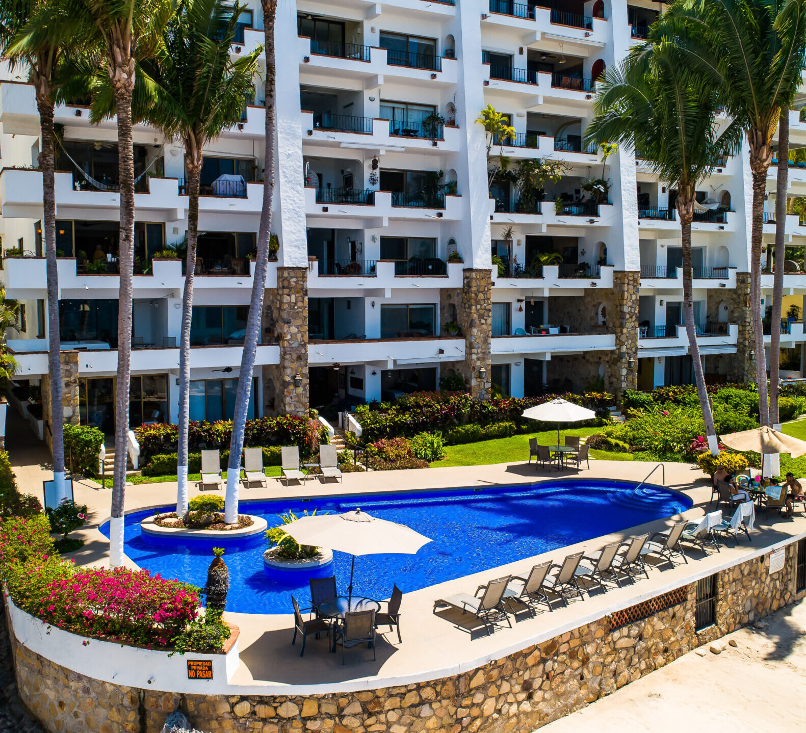 A view looking down at a curved blue swimming pool surrounded by palm trees in front of a white multi-story condominium building.